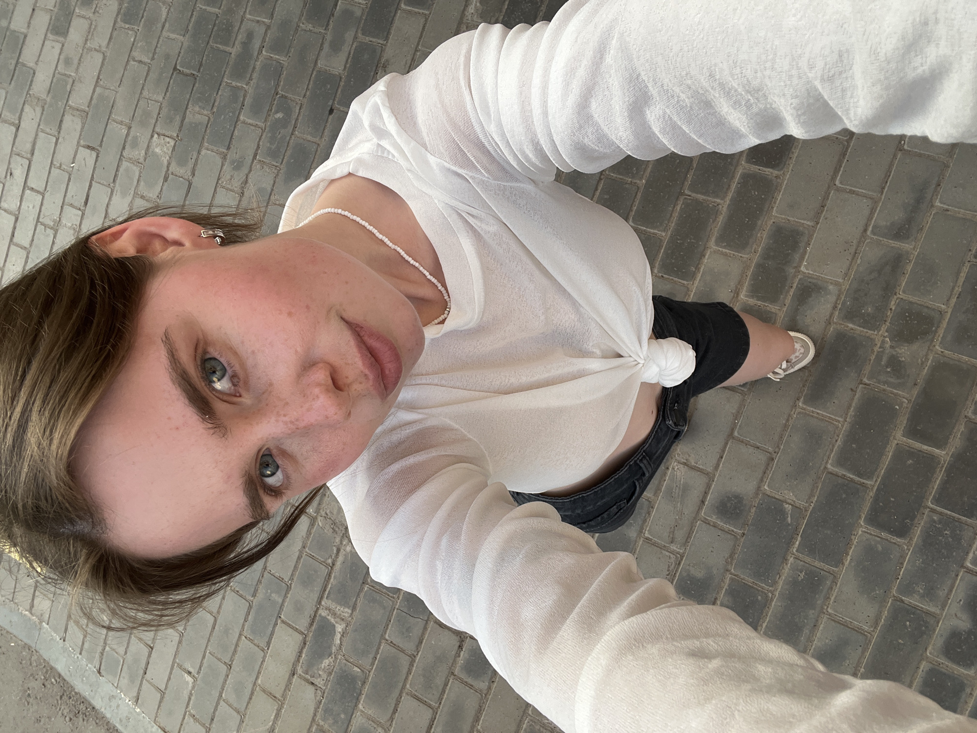 A high-angle upward-looking selfie captures a young adult female on a paved surface in the city of Chaikovsky, Russia. She has medium-brown hair, blue eyes, and a fair complexion with scattered freckles on her nose and cheeks. Her lips are slightly parted, and she looks directly at the camera. She is dressed in a sheer white long-sleeved top, knotted at the front to reveal her lower abdomen, dark denim shorts, and light-toned low-top sneakers visible on her right foot. Accessories include a white beaded choker necklace and small silver-toned hoop earrings. The ground is composed of dark grey rectangular brick pavers laid in a stretcher bond pattern, with a lighter grey asphalt or concrete surface visible along the upper-left edge of the frame.