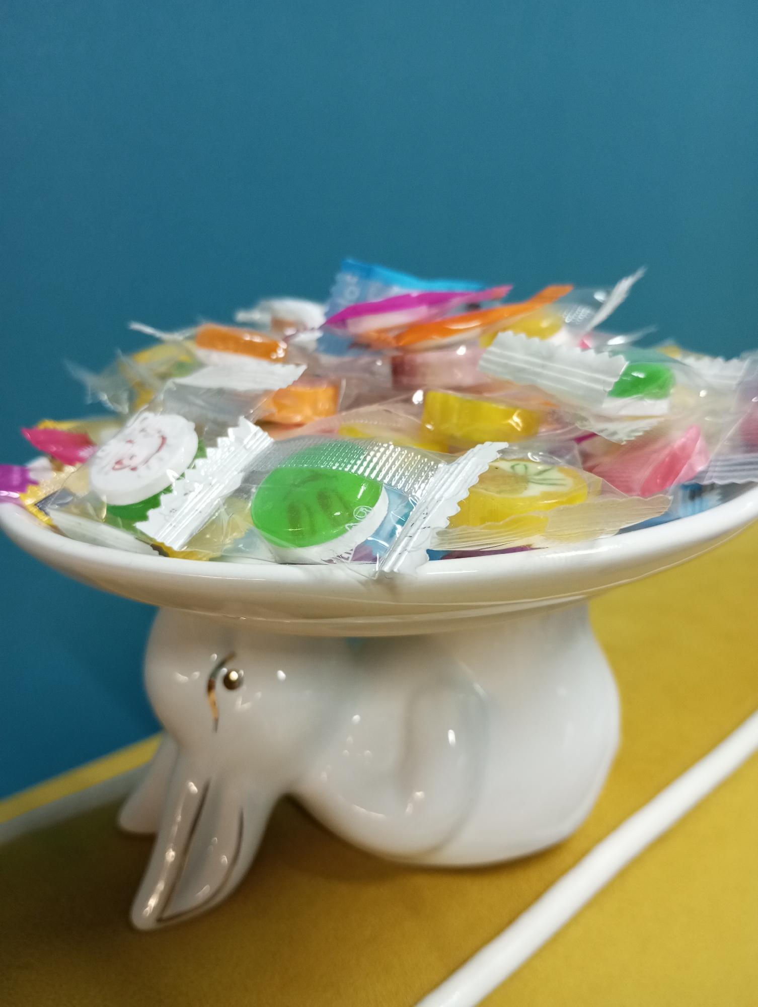 A close-up shows a white, ceramic elephant stand topped with a shallow, round dish overflowing with individually wrapped hard candies in various colors. The candies are mostly round and wrapped in clear plastic with crimped edges. The background is split between a blue wall above and a yellow surface below. The setting does not appear to be identifiable as Pervomaysky, Russia, from the provided image alone.