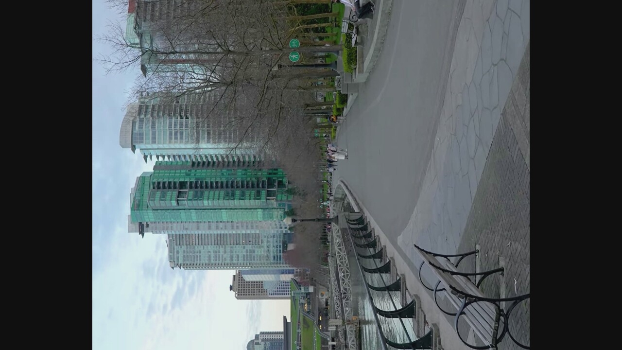 This image depicts a scenic urban waterfront in Vancouver, Canada, likely False Creek, with modern high-rise buildings forming the backdrop. The foreground features a paved walkway lined with mature trees that have lost their leaves, indicating a season other than summer. Several park benches are visible along the walkway, one occupied by a person.
In the middle ground, people are strolling along the path, enjoying the waterfront. A body of water, possibly a harbor or inlet, is to the left, with a decorative railing separating it from the walkway. Beyond the water, structures that appear to be docks or piers are visible.
The architecture of the buildings is contemporary, characterized by glass facades, some with a distinctive green tint. The sky is overcast, contributing to a diffused light that softens the scene. The overall impression is one of a well-maintained public space in a bustling city, offering a pleasant environment for recreation and sightseeing.