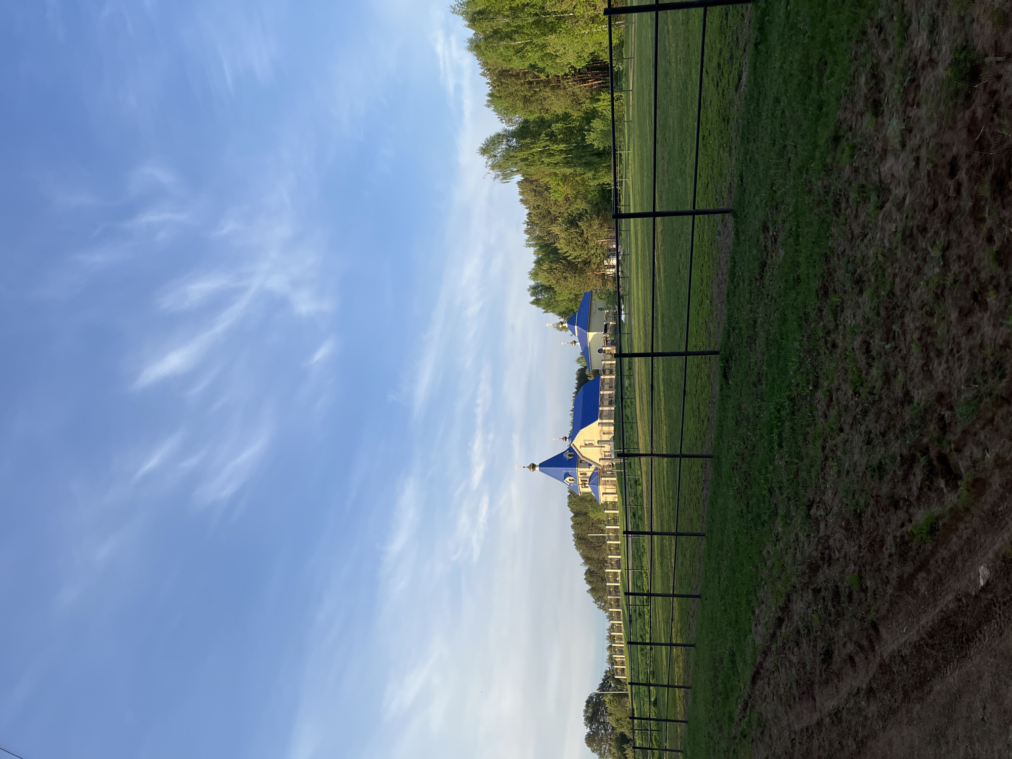 A photograph captures a scene featuring a building with distinctive blue domed roofs, indicative of a religious structure, likely a church. The building is situated on a grassy field, delineated by a dark, dirt-like area in the foreground and a line of trees in the background. A series of fence posts extends along the edge of the field, adding depth to the composition. The sky above is a pale blue, adorned with wispy clouds.
The photograph is of the Church of the Icon of the Mother of God "Joy of All Who Sorrow" in Chaikovsky, Russia.