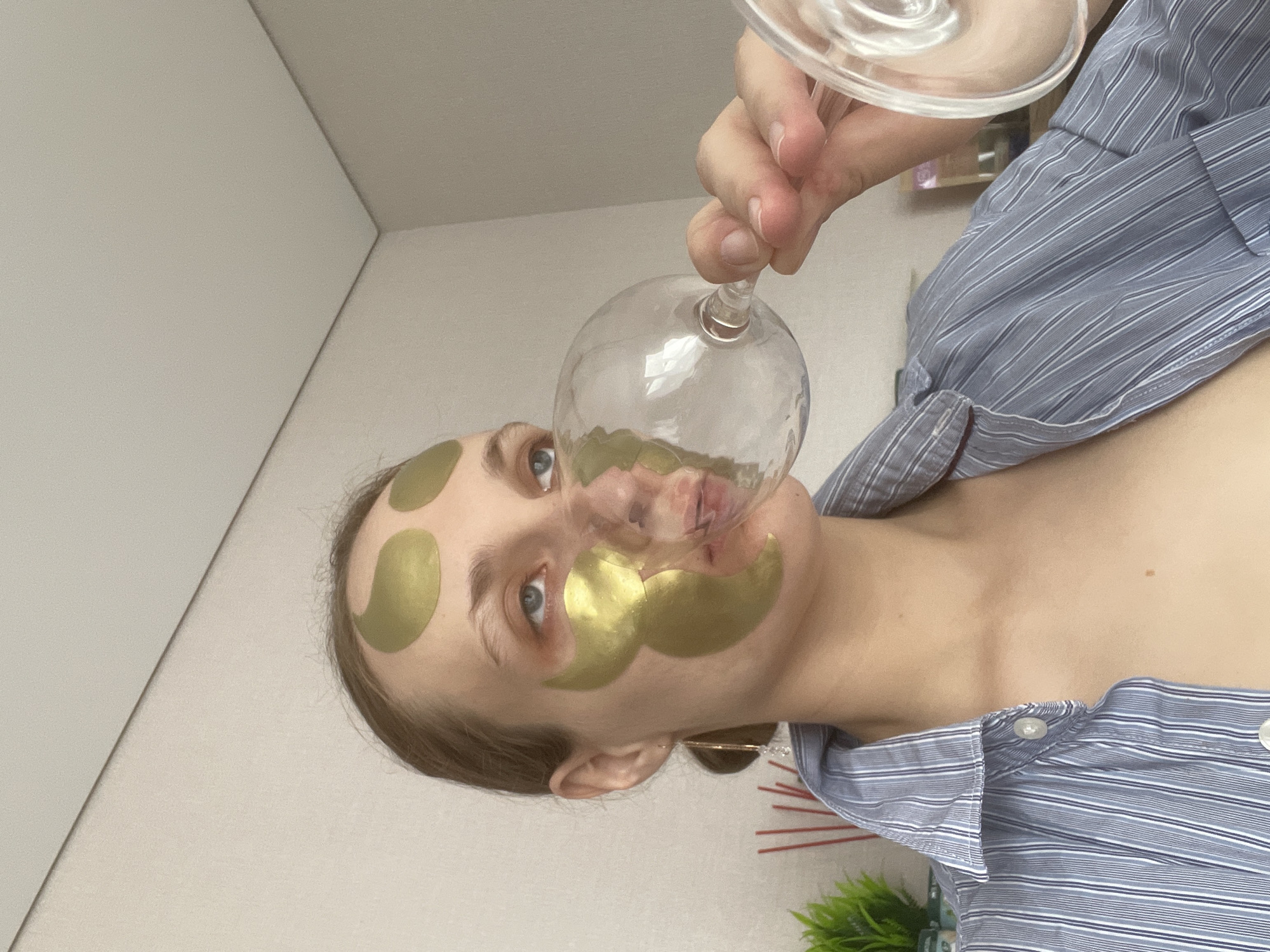 A woman with gold eye and forehead patches holds an upside-down wine glass to her face. She wears a blue-and-white striped shirt. The background includes a plain wall, ceiling, and a small green plant. It is not possible to determine if the scene is in Подшивалово, Russia, or any other specific location based solely on the visual information provided.