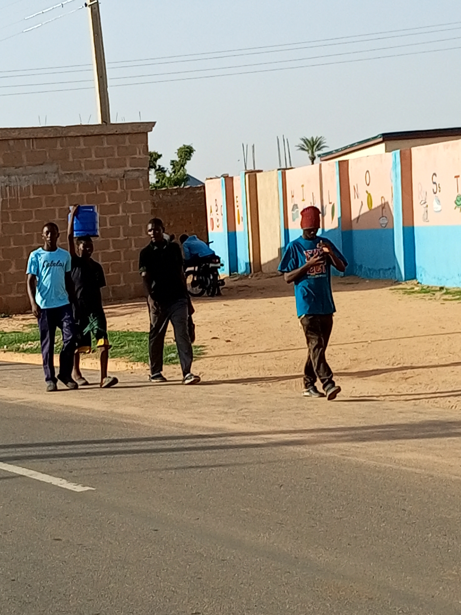 In Tempure, Nigeria, four young men walk on a dirt road adjacent to a paved road. On the left, two men walk side-by-side; the man on the left wears a light blue t-shirt and dark pants, while the man to his right wears a black t-shirt and green shorts. The man in the black t-shirt carries a large blue water jug on his head. To the right of these two, another man in a black t-shirt and dark pants walks alone. Further to the right, a fourth man, wearing a blue t-shirt and dark pants, walks with his head bowed. Behind the men, a colorful wall with letters and drawings of objects is visible, suggesting the presence of a school. A brick wall stands to the left of the scene. A motorcycle is parked in the background. The sky is clear and bright, indicating daytime.