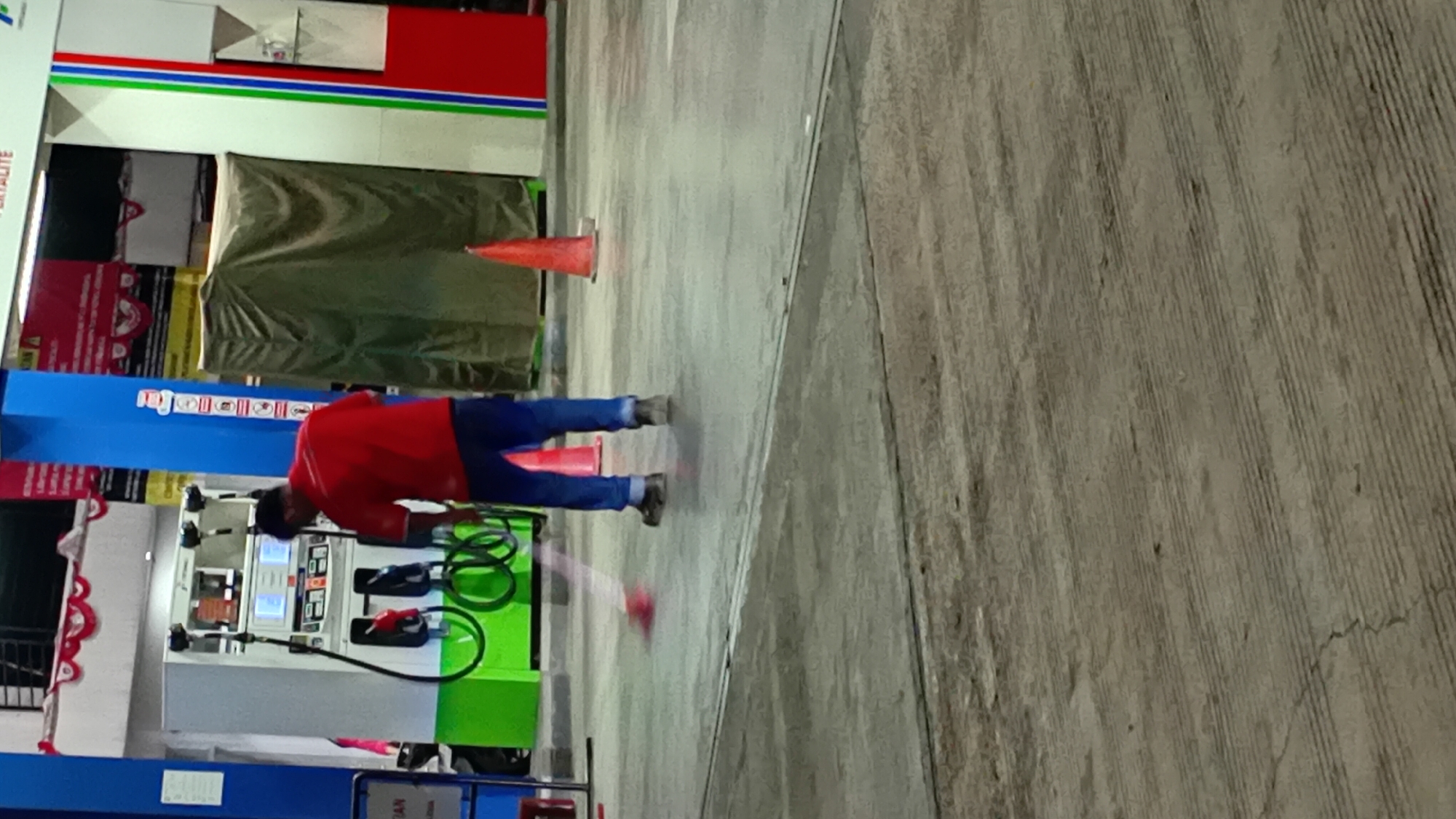 A person wearing a red shirt and blue pants is sweeping the concrete ground at a gas station in Kasang Pudak, Indonesia. The gas pump on the left has a green base and several hoses. In the background, a structure covered with a tan tarp is visible. Two orange traffic cones are on the ground. The scene is illuminated by artificial light.