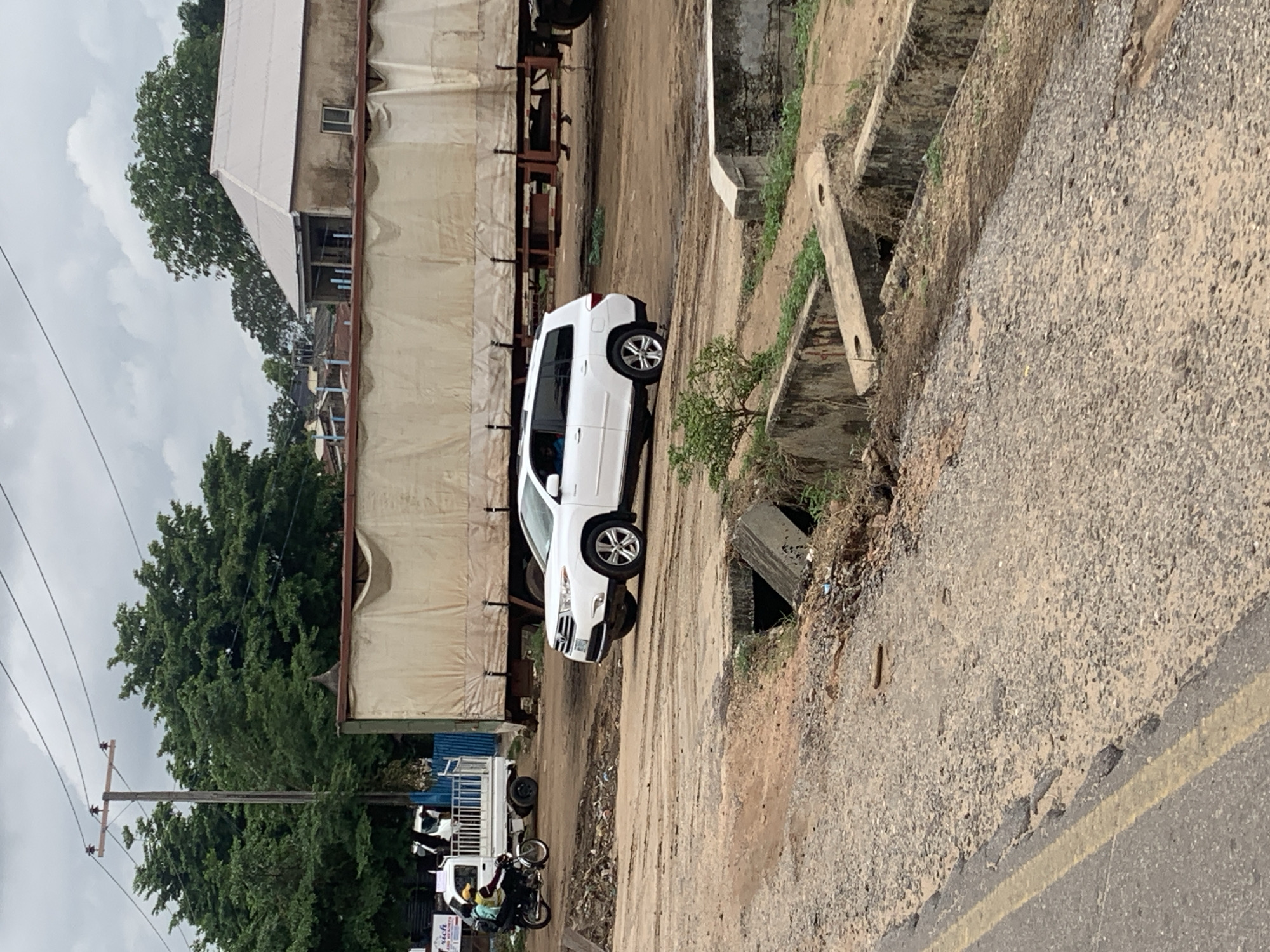 In Adankolo, Nigeria, a white Toyota Highlander SUV is navigating a bumpy, unpaved road, its front wheel appearing to be caught in a hole or on an uneven surface. The vehicle is positioned near a large, beige-covered trailer, suggesting a possible commercial area or transport hub. In the background, lush green trees contrast with the dusty road and a partially constructed building with a light-colored roof. A utility pole with power lines stands tall, and in the foreground, a concrete drainage structure with openings is visible. A motorcycle with two individuals is seen in the middle distance to the left, moving away from the camera. The sky is overcast, indicating a cloudy day.