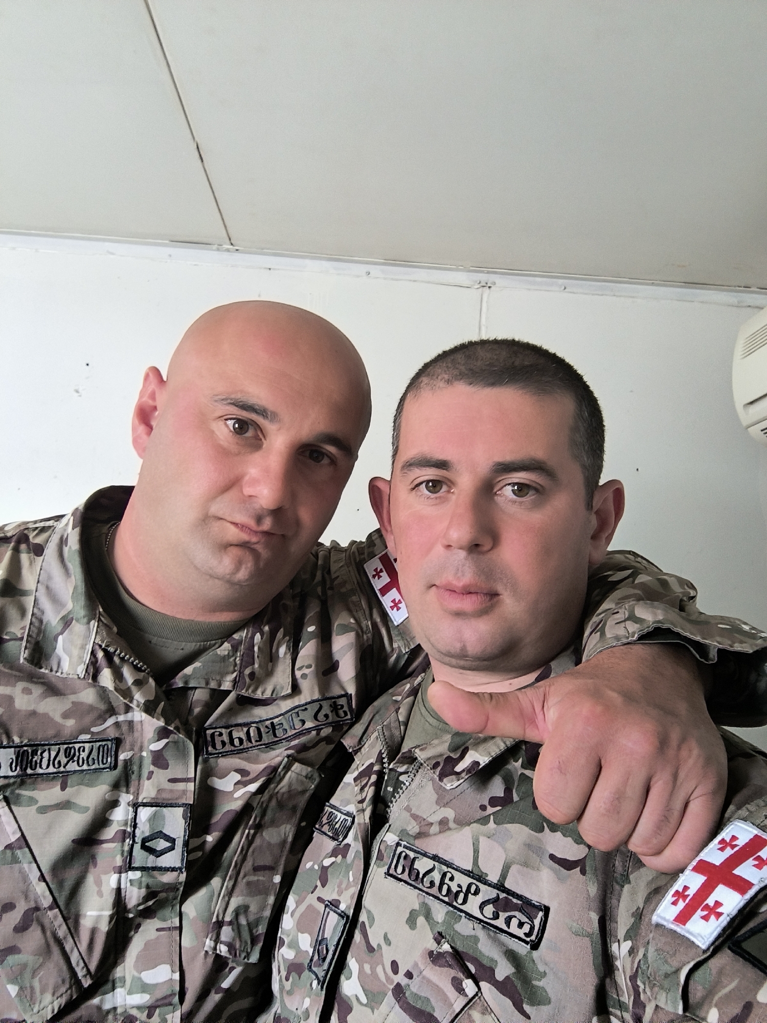 Two men are standing close together, wearing military camouflage uniforms with Georgian flags on their sleeves. The man on the left is bald and has a neutral expression. The man on the right has short hair and his hand is raised in a thumbs-up gesture. The background is a white wall. Tbilisi, Georgia is not discernible from the scene details.