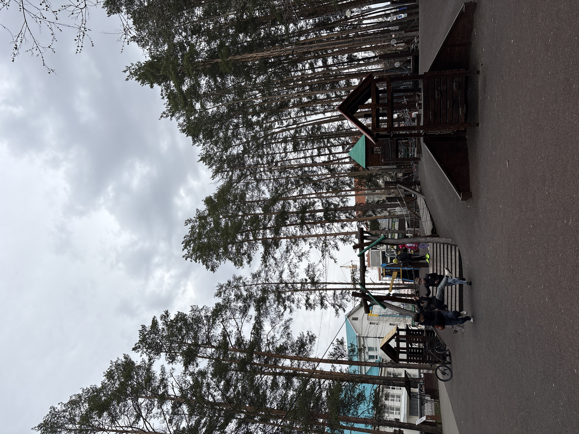 This media file captures an outdoor scene, likely a park or recreational area in Neftekamsk, Russia, on an overcast day. The dominant feature is a dense line of tall, slender conifer trees with dark green foliage, extending upwards and bordering the paved ground on the left. The sky above is uniformly grey with visible clouds, indicating a cloudy or overcast weather condition.
In the mid-ground, a wooden playground complex is visible. It features a main structure with a dark reddish-brown gabled roof and a distinct smaller section with a bright green triangular roof. Adjacent to it is another wooden play structure with a slide and climbing elements, identifiable by its green support bars. A dark-colored bicycle is parked on the dark, paved ground, which appears to be asphalt.
Several people are present, engaged in casual activities. Two young individuals, dressed in dark jackets and jeans, stand on the paved area near the play structures, seemingly socializing. Further back, on a raised wooden platform or steps of the play area, a group of people, including children, are gathered. Some wear brightly colored attire, such as a pink hat and a yellow jacket.
In the background, behind the trees and play area, residential or commercial buildings are visible. One light-colored building is partially covered with blue mesh or scaffolding, suggesting ongoing construction or renovation. The overall mood is casual and relaxed, indicative of a typical day at a community recreational space. There is no discernible text visible in the image.
