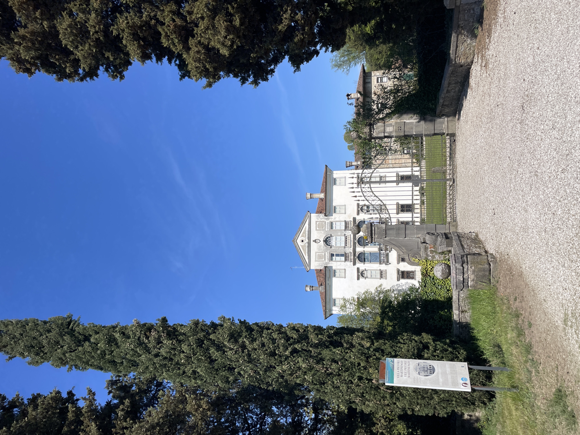 The image is an eye-level shot of a white, multi-story building in San Lorenzo, Italy. The building, partially obscured by trees and an iron gate, features architectural details such as arched windows, a triangular pediment, and red tile roofs. The sky is a clear, deep blue. Tall, dark green cypress trees frame the building on either side. In the foreground, a gravel path leads towards the gate. A sign with text and an image is visible on the right side of the path.