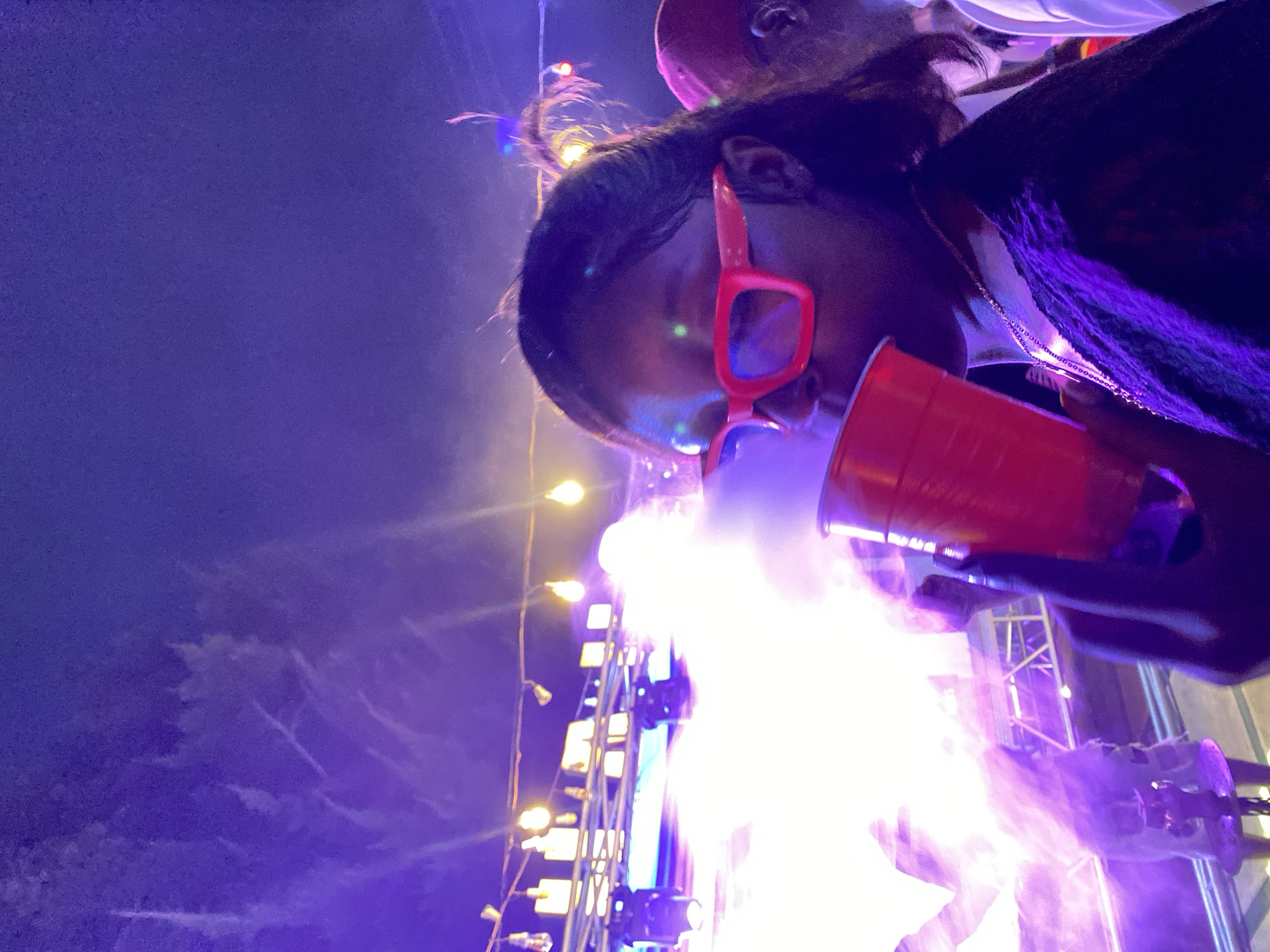 This is a medium shot, taken at night, of a young Black woman wearing bright pink square-framed glasses and a black top, holding a red plastic cup to her lips. She is looking directly at the camera, her expression neutral. Smoke or vapor is rising from the cup, obscuring her face and some of the background. The scene appears to be an outdoor event, likely a party or concert, illuminated by bright purple and white lights and some yellow string lights. In the background, other people are visible, as well as stage lighting and equipment, suggesting a lively atmosphere. The location is identified as Gura Top, Nigeria.