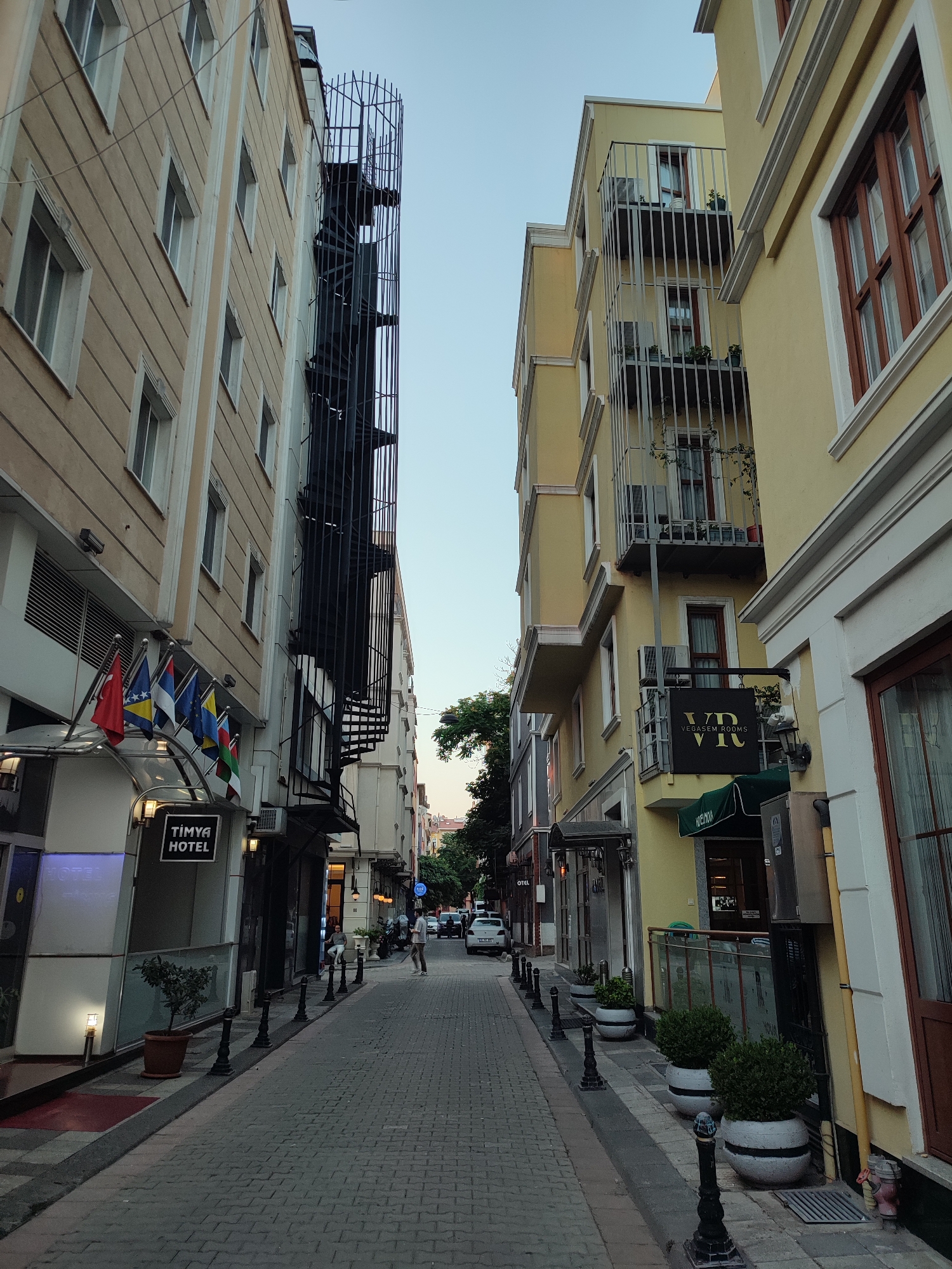 This is a street-level, eye-level, vertical shot of a narrow cobblestone street in Kadıköy, Turkey, with buildings lining both sides. The sky is clear and light blue. On the left, a multi-story building features a dark spiral staircase attached to its exterior, with a sign for "TİMYA HOTEL" and several flags displayed above the entrance. The building itself is light beige with white trim and numerous rectangular windows.
On the right, a yellow-painted building has multiple balconies with decorative metal railings, some containing potted plants and outdoor seating. A sign for "VR VEGASRM ROOMS" is visible. This building also has prominent wooden-framed windows.
In the middle distance, a few vehicles are parked or moving slowly down the street. Two individuals are visible: one standing near the hotel entrance on the left, and another standing further down the street on the right, facing away from the camera. Small potted plants are placed along the edge of the sidewalk on the right side of the street. The overall impression is a charming, somewhat historical urban street scene.