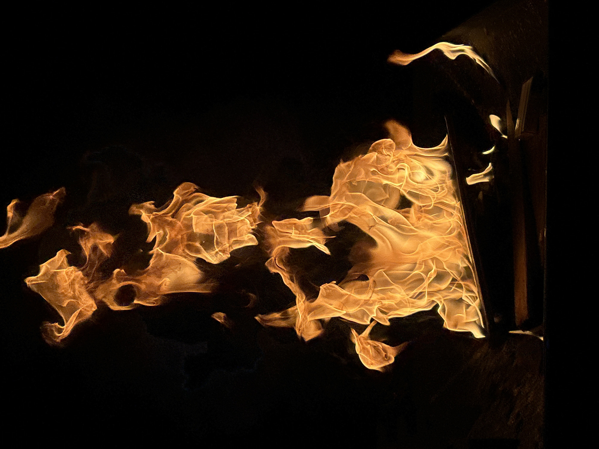 The image displays a close-up, vertical shot of a vibrant bonfire against a completely black background. The flames, rendered in brilliant shades of orange and yellow, are the sole subject of the photograph, dominating the frame with their dynamic shapes and intense luminosity. They appear to rise from a dark, indistinct object that could be logs or a fire pit, situated at the bottom of the image. The flames exhibit a fluid, dancing motion, with wisps and tendrils reaching upwards, creating a sense of movement and heat. The overall impression is one of raw, natural energy. There are no people, animals, or any other discernible objects or environmental features visible in the image. The setting is entirely abstract due to the black background, offering no clues about its specific location. Therefore, it is impossible to determine if this scene is in Chaikovsky, Russia, or any other specific place.