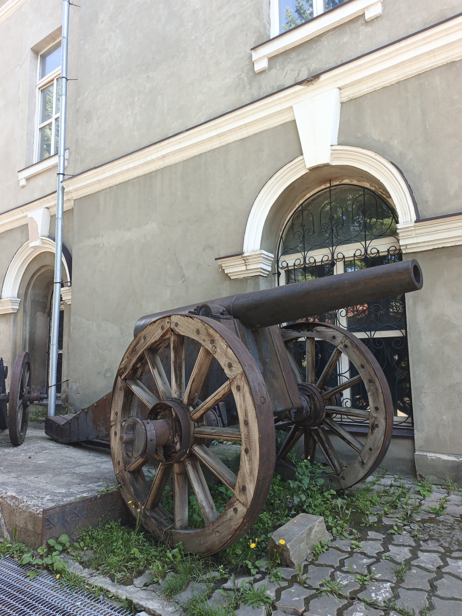 An old cannon is on display outside a grey building in Zlatoust, Russia. The cannon, made of rusted metal and wood, has large spoked wheels. The building has a light grey facade with white trim around the windows and arched doorways. One of the arched doorways has a decorative wrought-iron gate visible behind the glass. Green grass and weeds grow around the base of the cannon and between the patterned stone pavers on the ground. A metal downspout runs vertically along the side of the building.
