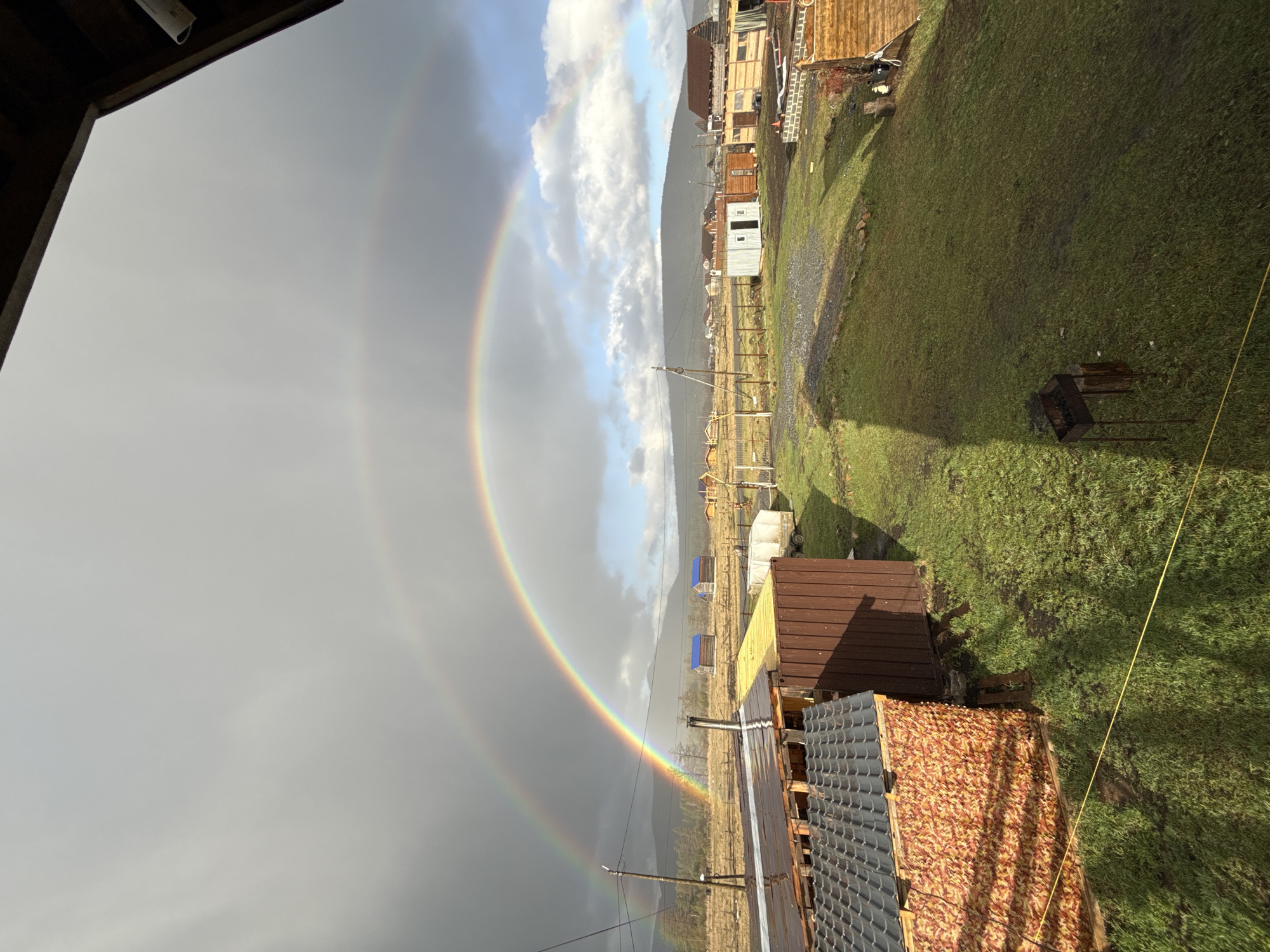 This image captures a vibrant double rainbow arching over a rural or semi-rural settlement, likely in Tyulyuk, Russia, after a rain shower. The scene is viewed from a slightly elevated position, possibly from a window or balcony.
**Subjects:** The primary subject is a striking double rainbow, prominently displayed against a dramatic sky. The scene also features a collection of modest buildings, green landscape, and distant mountains. No people are visible in the image.
**Scene and Setting:** The setting appears to be a small village or cluster of homes nestled in a valley or at the base of hills. The ground is covered in lush green grass, suggesting recent rainfall and fertile land. Dirt tracks or unpaved paths crisscross the area. Hazy mountains form the backdrop in the distance.
**Activity or event:** The main event captured is the natural phenomenon of a double rainbow appearing after a period of rain. There is no visible human activity, lending a peaceful and serene atmosphere to the scene.
**Notable details:**
* **Time of day and weather:** The presence of the rainbow, along with the mixed sky, indicates a recent rain shower followed by clearing conditions. One side of the sky is dominated by dark grey clouds, while the other shows patches of blue sky and bright white clouds, suggesting the sun is shining from behind the viewer. The lighting suggests either late afternoon or early morning.
* **Rainbow:** A bright, distinct primary rainbow is clearly visible, with a fainter, larger secondary rainbow arching above it.
* **Buildings:** Several rustic buildings are scattered across the landscape. In the foreground, part of a dark brown corrugated metal roof is visible, with a section featuring a unique orange and red patterned material. Further back, wooden houses and smaller sheds, some with light-colored siding, are present. A small, square white structure resembles an outhouse or utility shed.
* **Objects:** A metal barbecue grill (mangal) sits on the grass in the mid-ground. Power poles and overhead wires traverse the scene. A yellow rope or string stretches across the lower right portion of the image.
* **Location cues:** The general rustic appearance of the buildings and the mountainous landscape are consistent with the provided location context of Tyulyuk, Russia.
**Visible text:** No visible text is present in the image.
