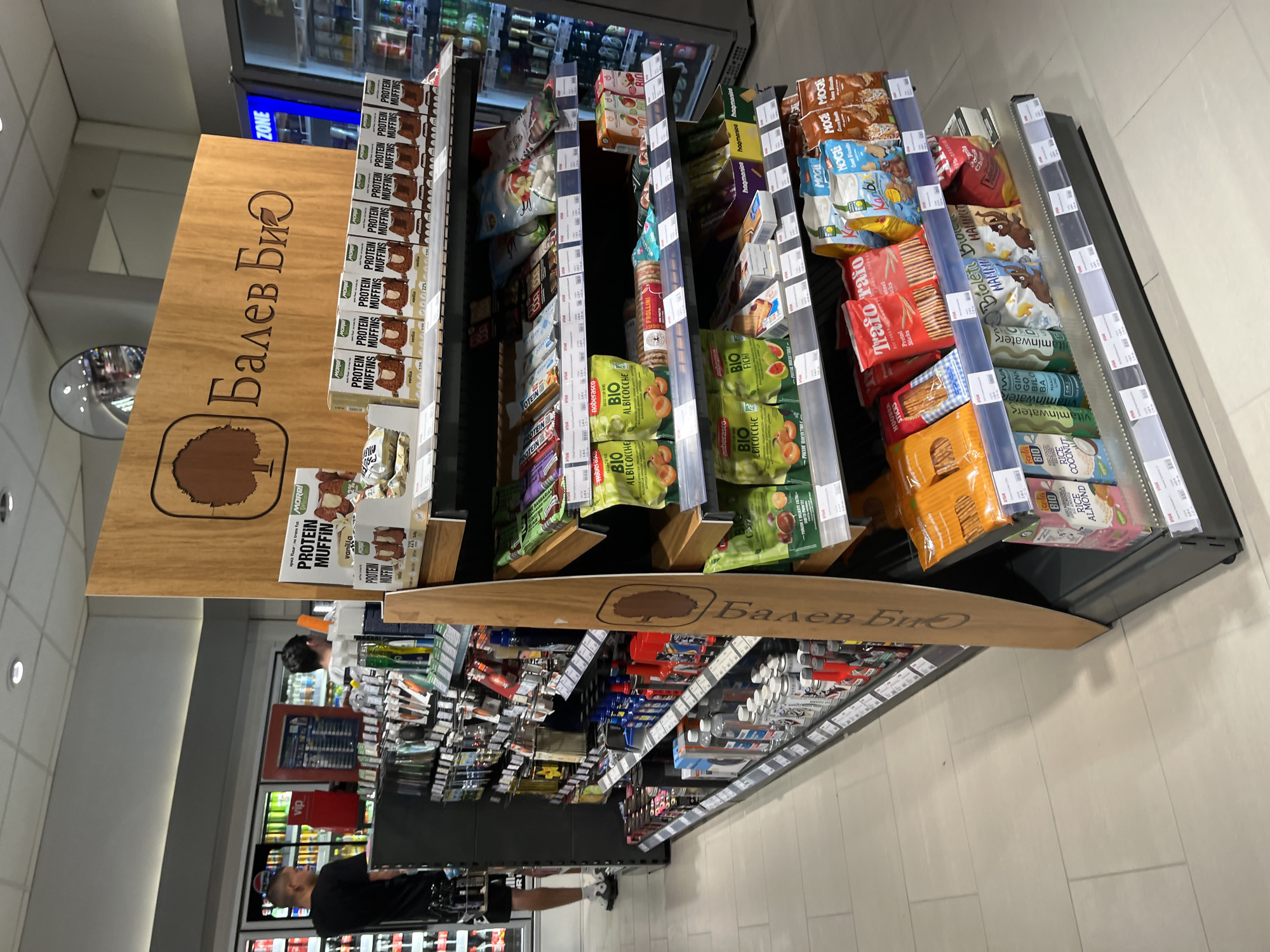 The image shows a shelf in a shop with a variety of snacks and health foods. The shelf is labeled "Балев Био," which is written in Cyrillic script. There are several different brands and types of products, including protein muffins, dried fruit, and crackers. There is a man with a black shirt in front of the shelf. The bottom part of the image shows more shelves with groceries. The location of the shop is a shopping area, possibly a supermarket. There is no visible indication that the shop is in the city of Popovitsa, Bulgaria.