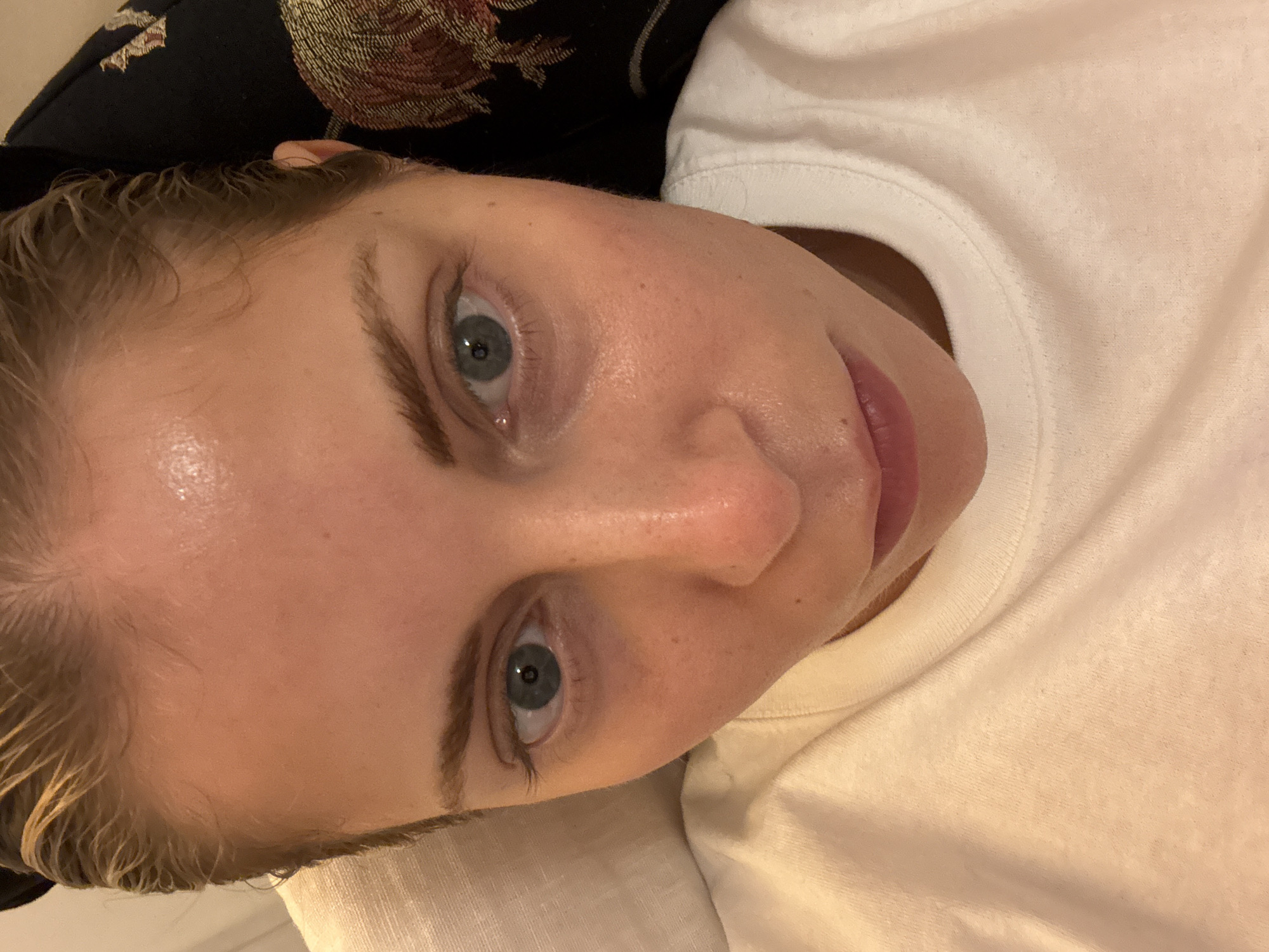 A close-up shot of a person's face. The person has fair skin, light eyebrows, and blue eyes. The hair appears to be blonde and slightly wet. The person is wearing a white t-shirt. The background consists of a dark patterned fabric and a white pillow. The image does not show any indication of being in Paris, France.