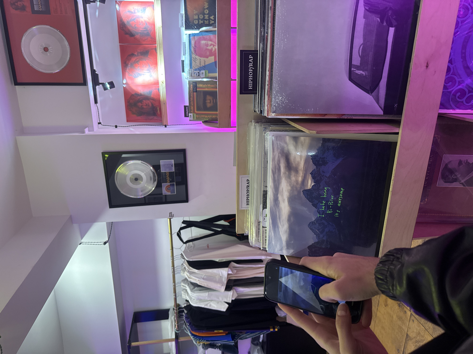 A person is holding a smartphone, taking a photo of a vinyl record in a shop. The record's cover features a landscape with mountains, and someone has written on the cover "I hate being Bi-Polar, it's awesome" in green marker. The album is in the hip-hop/rap section. Behind it are other records and a neon purple light. To the left of the albums is a clothing rack with shirts. Above the albums are framed music awards. There are framed silver records hung on the wall. Based on the record covers, the shop appears to be located in Yerevan, Armenia.