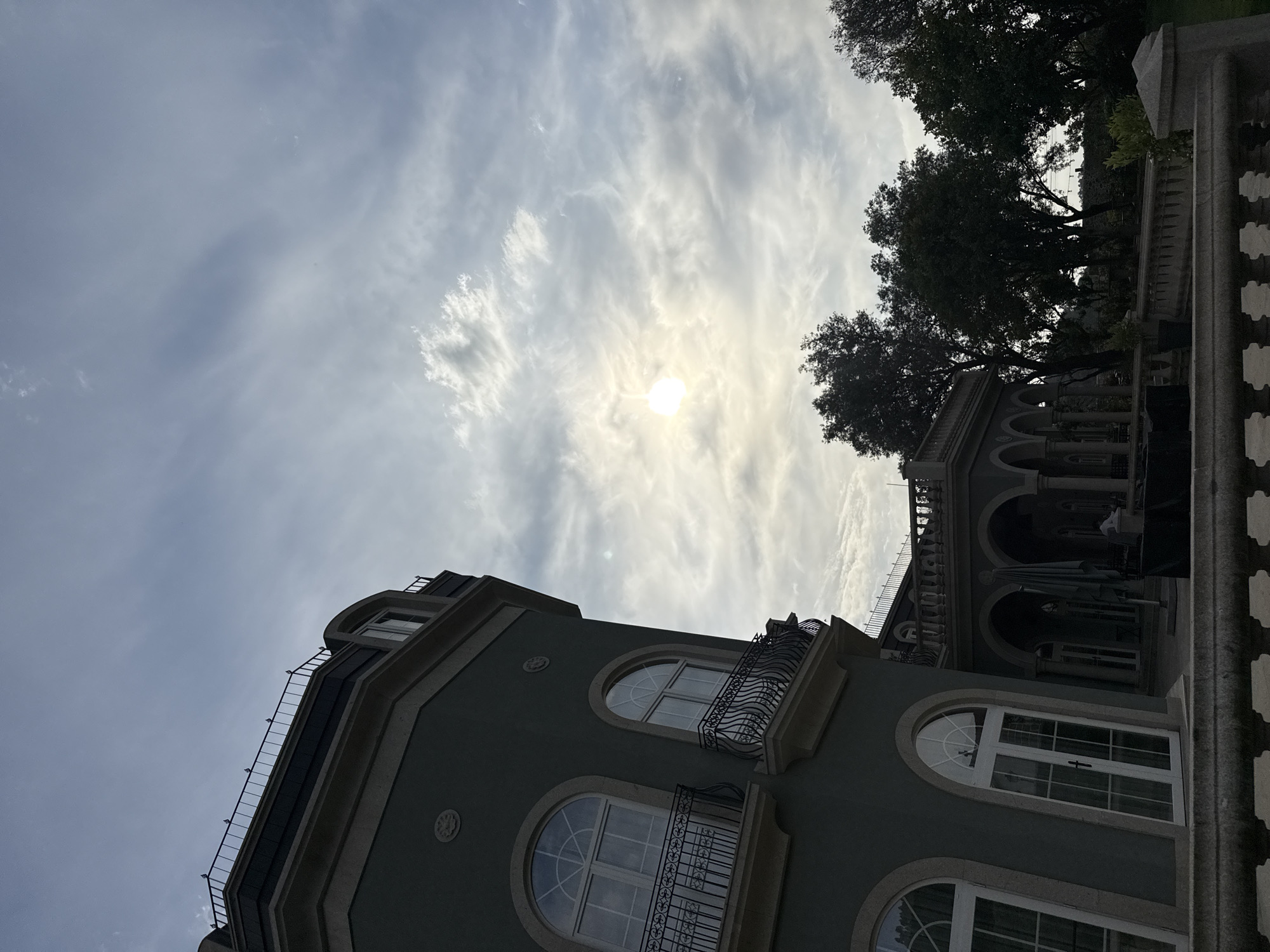 The image is a low-angle shot of a building in Vila Verde, Portugal. The building is multi-story, with arched windows and balconies. The exterior is a muted gray color. The upper portion of the frame is filled with a sky of light, wispy clouds, with the sun shining faintly through. To the right, there are dark silhouetted trees and a stone balustrade. The scene appears to be taken during daylight hours, possibly late afternoon, given the direction and diffusion of the light.
