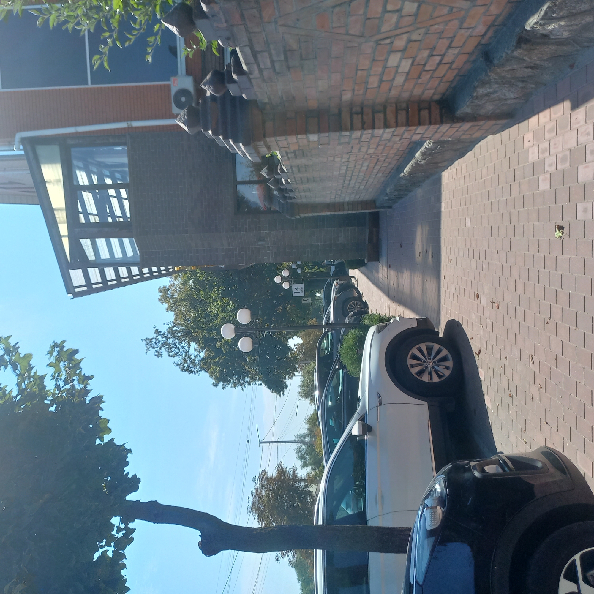 The image captures a sunlit street scene in Kropyvnytskyi, Ukraine, likely in front of a cafe identified by a sign with the number "14" and the word "coffee." In the foreground, a black car is partially visible on the left, and a white SUV is parked next to it. The pavement is paved with reddish-brown bricks, and a shadow from a tree stretches across it.
To the right, a brick building stands with a dark grey brick facade and a multi-story window structure with a glass roof. An air conditioning unit is mounted on the brick wall. A decorative brick fence with pointed stone elements runs along the side of the building.
Further down the street, a series of three-globe streetlights are visible, and another similar streetlight is positioned near the "14 coffee" sign. Trees with green foliage line the street and the background. The sky is clear and blue. There are no people visible in the image. The overall impression is one of a well-maintained, modern urban environment.