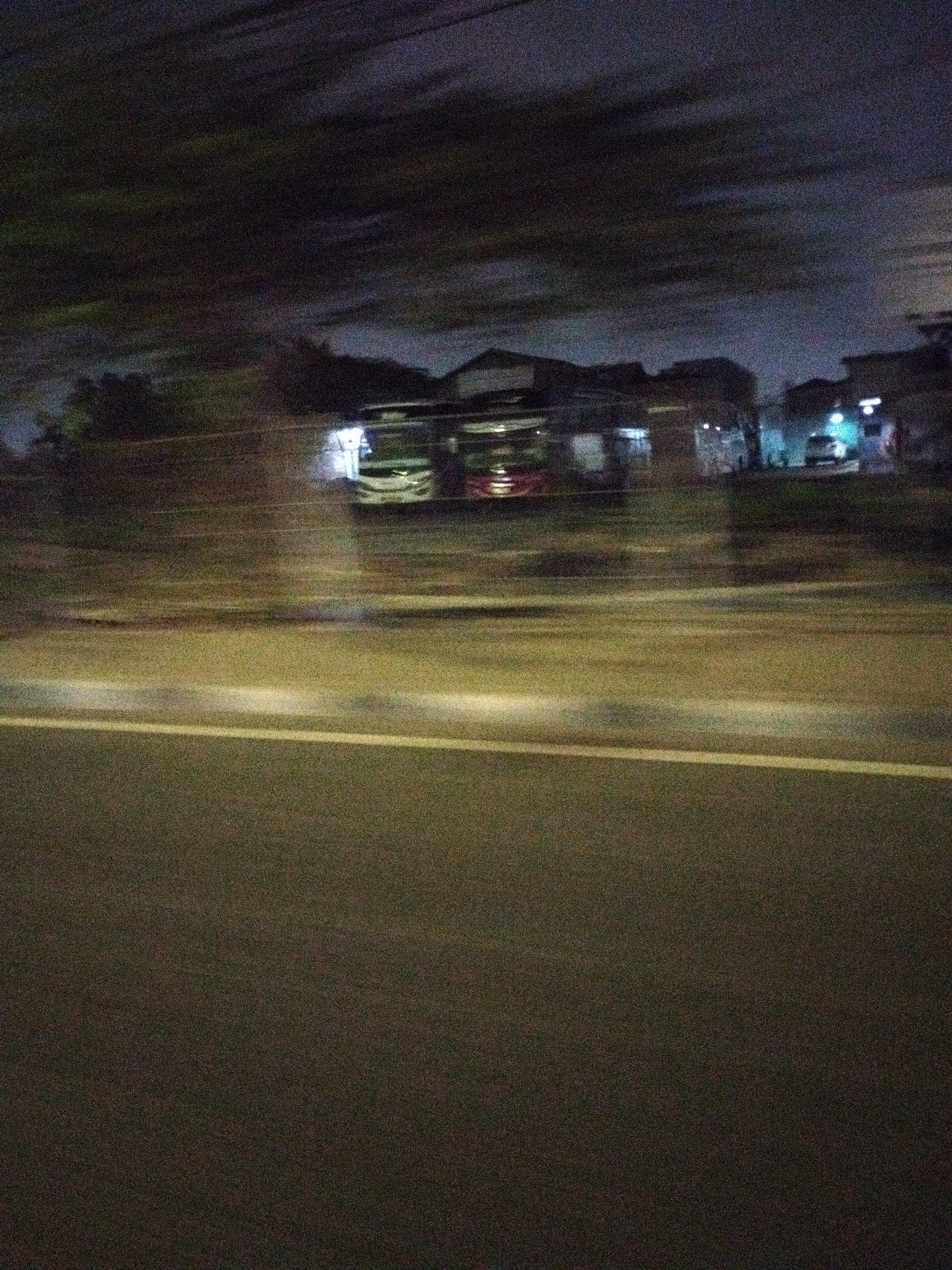The image depicts a blurry, nighttime scene with a motion blur effect, suggesting it was taken from a moving vehicle. The foreground is dominated by a dark asphalt road with a white line dividing the lanes. In the middle ground, several buildings are visible, appearing as silhouettes against a dark sky. Two buses, one with red accents and the other white, are parked in front of one of the buildings. A smaller white vehicle is also parked further down. The lighting suggests artificial illumination from streetlights or building lights, casting faint glows and reflections. The overall impression is one of a street scene in an urban area at night, likely in Surabaya, Indonesia, given the context.
