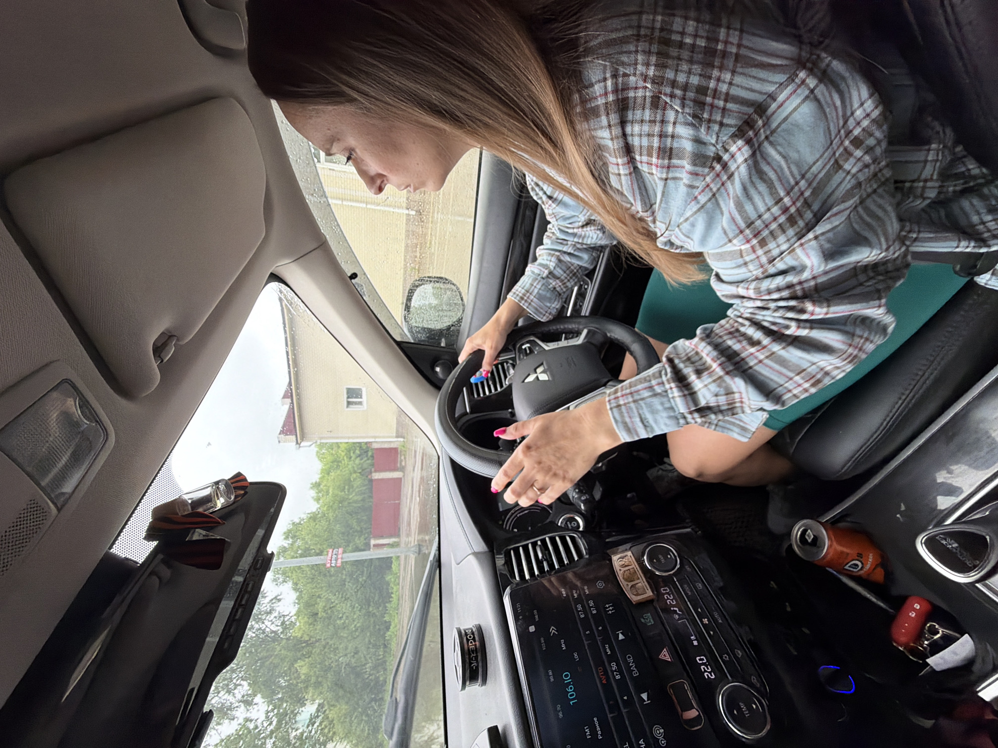 A woman drives a car through the city of Birobidzhan, Russia, under overcast skies. Rain streaks the windshield, and puddles are visible on the road. She is wearing a plaid shirt and green skirt. Her hands are on the steering wheel, and her fingernails are painted bright pink. A can of orange soda sits in the cup holder, along with a set of keys. The car's dashboard display shows the temperature is 22 degrees Celsius.