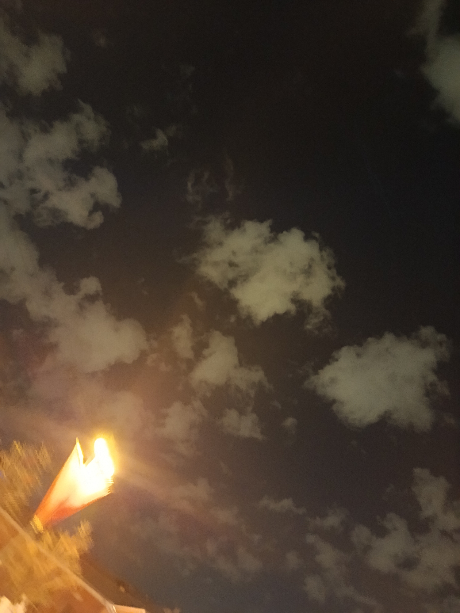 The image shows a night sky with scattered clouds. A bright light source, possibly a street lamp, is visible in the lower-left corner. Buildings and trees are partially visible and blurred in the foreground. It is not possible to determine the specific location in Moscow or any details about people, subjects, objects, actions, and interactions without additional information.