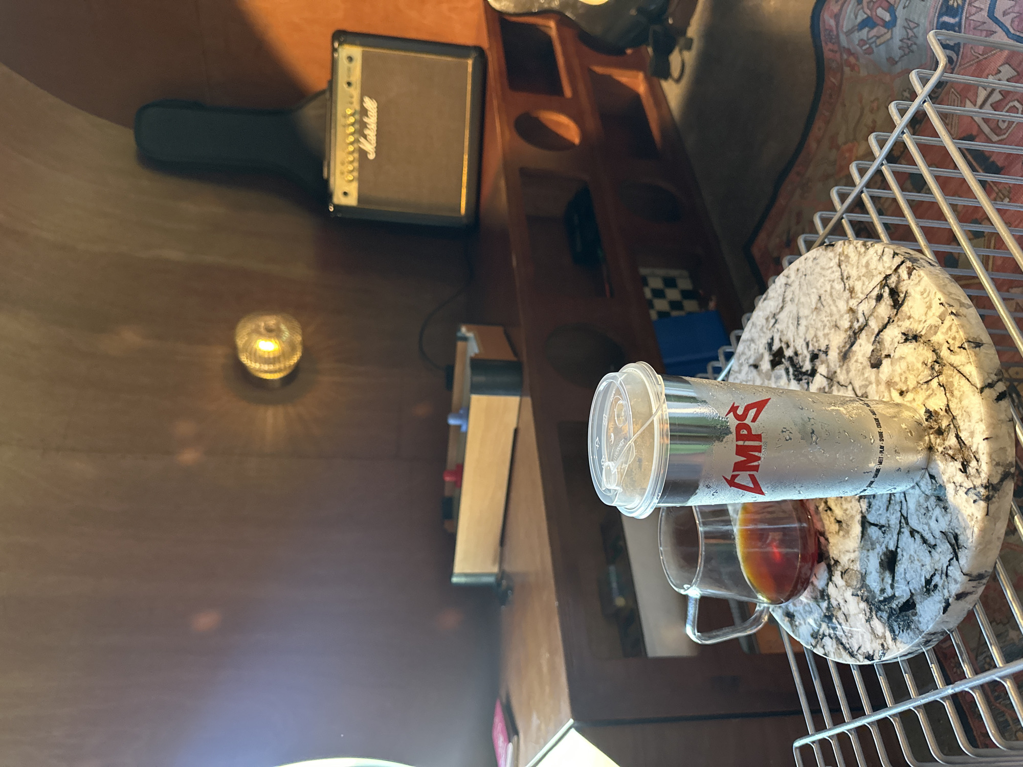The image shows a close-up view of a table with two drinks and a foosball game on a shelf in the background. In the foreground, a clear glass mug filled with dark liquid, likely coffee or tea, sits on a circular marble coaster. Next to it is a tall, silver, insulated cup with a clear plastic lid, condensation visible on its surface, and the red logo "CMPS" printed on it. Below the drinks, a wire rack is visible. In the midground, a wooden shelf holds a miniature foosball game with red and blue players. Behind the shelf, a Marshall guitar amplifier is visible, with a black guitar case leaning against the wall above it. A wall-mounted light fixture with a yellow glow is also present on the wooden wall. The overall scene appears to be indoors, possibly in a room with wooden paneling.