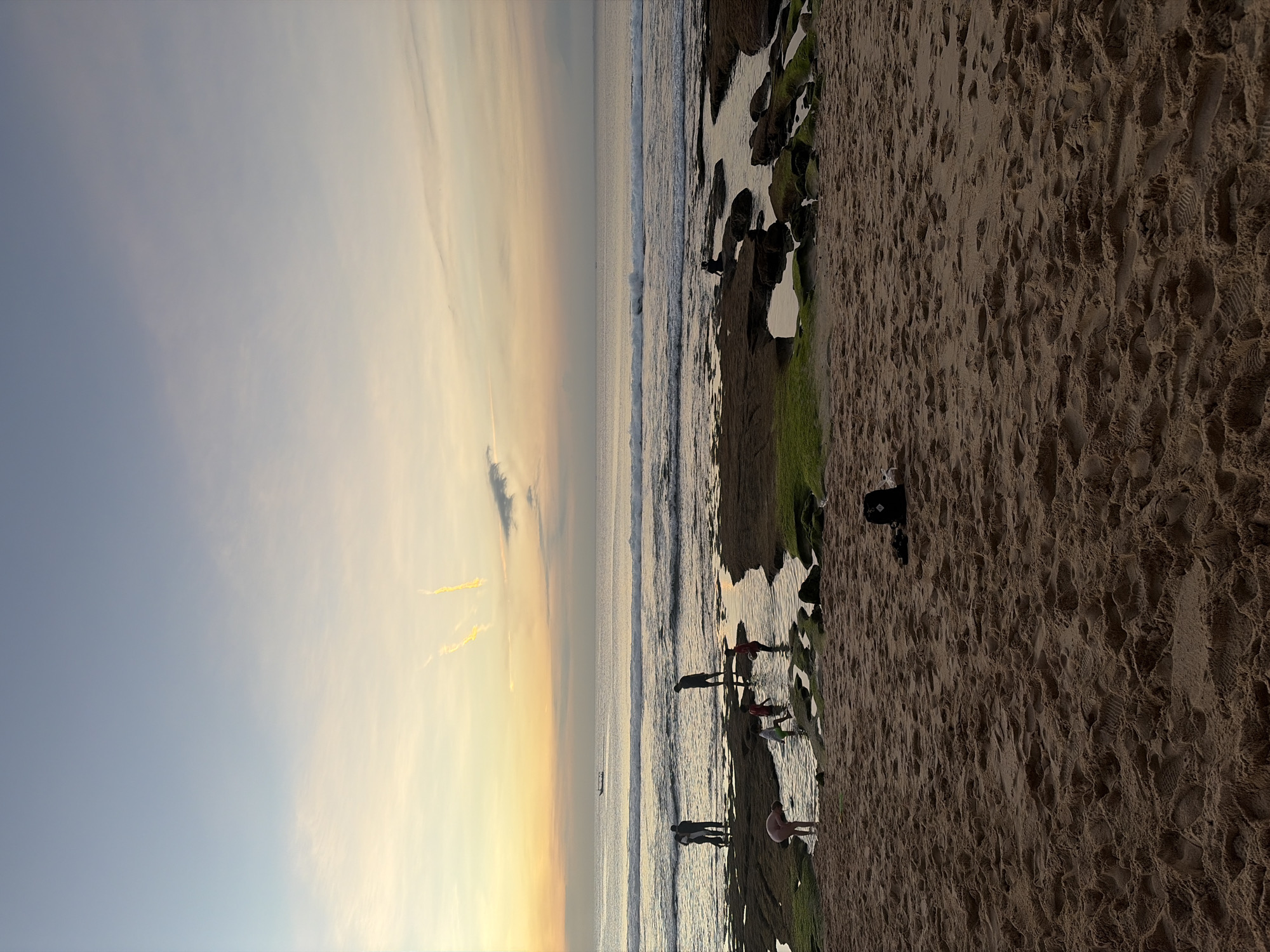 The image depicts a beach scene at dusk. The sky is a gradient of pale blue and soft yellow, with wispy clouds and two distinct yellow contrails high above. The ocean is calm with gentle waves breaking on a rocky shoreline. Several people are visible in the shallow water and on the rocks, some appearing to be children playing and adults observing. In the foreground, the sand is a warm brown, textured with footprints and ripples. A black backpack sits on the sand in the middle ground. A small boat is visible on the horizon. The scene conveys a peaceful and relaxed atmosphere.