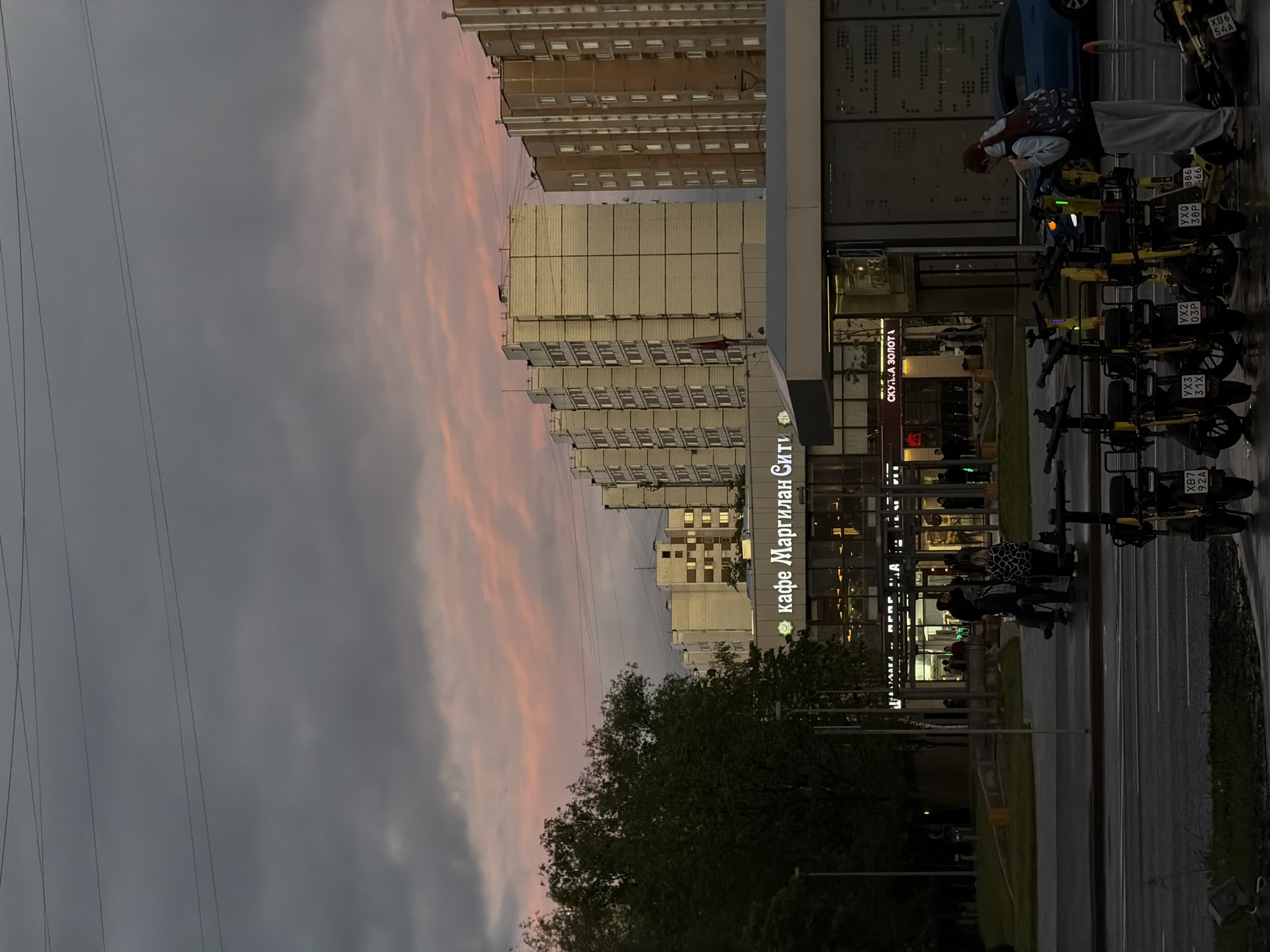 Here is a detailed description of the image:
**Overall Impression:**
The image presents a sideways view of an urban scene, likely taken in the evening or twilight. The sky is a mix of gray clouds and pink hues, suggesting either dusk or dawn. The focus is on a street scene with buildings, pedestrians, parked scooters, and a commercial establishment.
**Setting:**
* **Location:** Based on the signage, specifically the phrase "кафе Маргилан Сити", the photo is likely taken in Moscow, Russia. "Маргилан Сити" may refer to a specific area or a commercial establishment.
* **Environment:** The area appears to be a modern, urban environment. There are tall apartment or office buildings in the background. There are trees lining the street in the foreground.
**Objects and Structures:**
* **Buildings:** Several tall buildings are visible in the background.
* **Street and Sidewalk:** The photo shows part of a street with scooters and a sidewalk with pedestrians.
* **Transportation:** There are parked scooters lined up along the street, with license plates indicating their use for a ride-sharing program.
* **Commercial Establishment:** The building with the sign "кафе Маргилан Сити" (Cafe Margilan City) is prominently featured, indicating a commercial establishment.
**People:**
* **Pedestrians:** There are people visible on the sidewalk near the cafe. Some are standing around the parked scooters.
**Actions and Interactions:**
* People are walking and gathering near the commercial establishment.
* Some people may be interacting with the parked scooters.
**Lighting and Atmosphere:**
* The image appears to be taken during twilight or evening, as indicated by the sky and the presence of artificial lights.
* The overall mood is somewhat subdued and urban.
**Other Details:**
* Power lines are visible in the sky above the buildings.
* There is a sign on the left side of the frame with the words “СКУПКА ЗОЛОТА,” which means "gold buying."
**In Summary:**
The image depicts a street scene in Moscow, Russia, near a commercial area. The focus is on people and scooters near a Cafe Margilan City during twilight, with a backdrop of urban infrastructure.