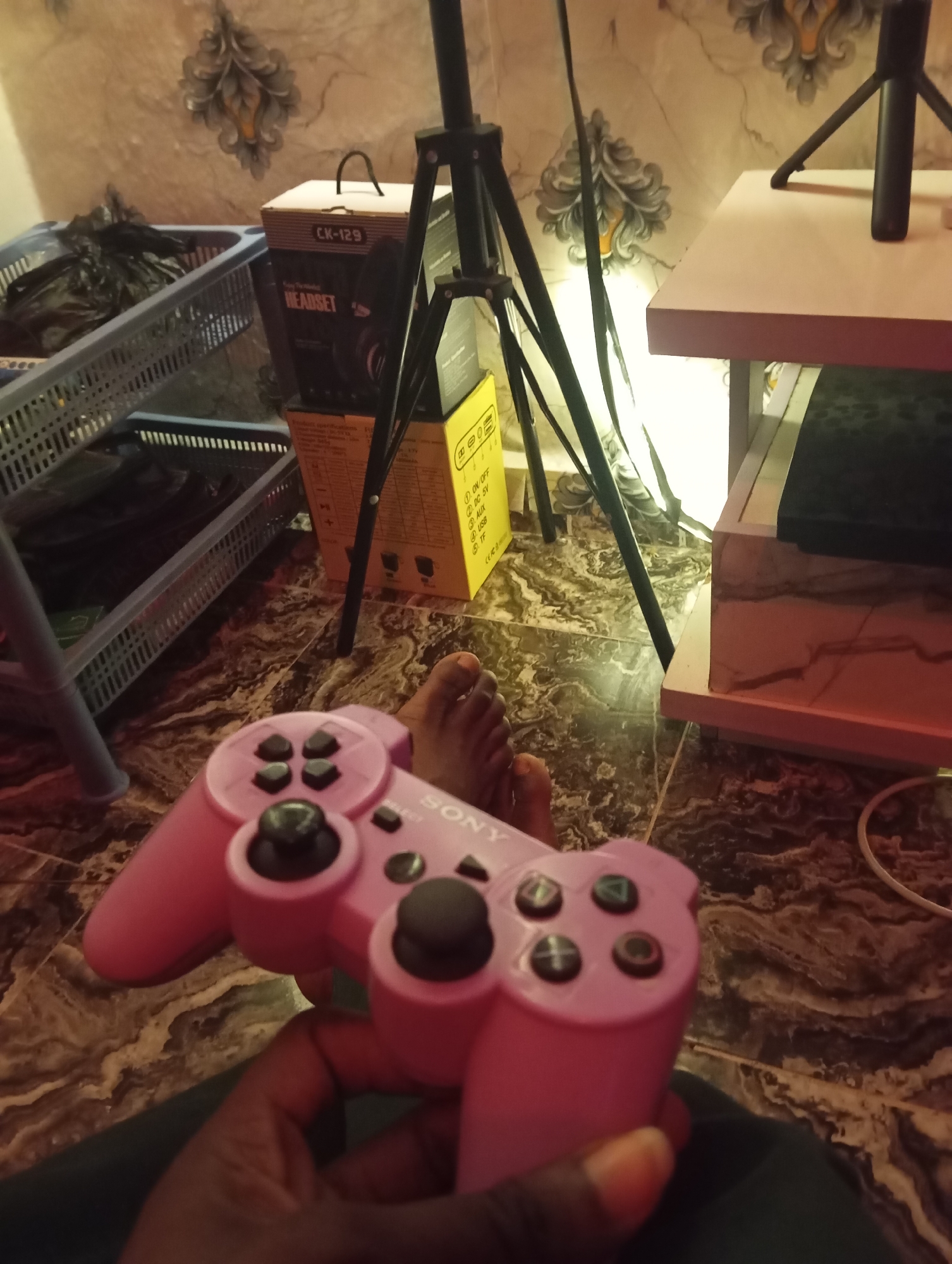 In Jos, Nigeria, a person's hand holds a pink video game controller, identified by the "SONY" logo. The controller is positioned in the foreground, with its buttons and joysticks visible. In the background, a black tripod with a light is set up near a shelf holding various items, including a box labeled "CK-129 HEADSET" and a blue storage basket filled with black bags. The floor is tiled with a marbled pattern.