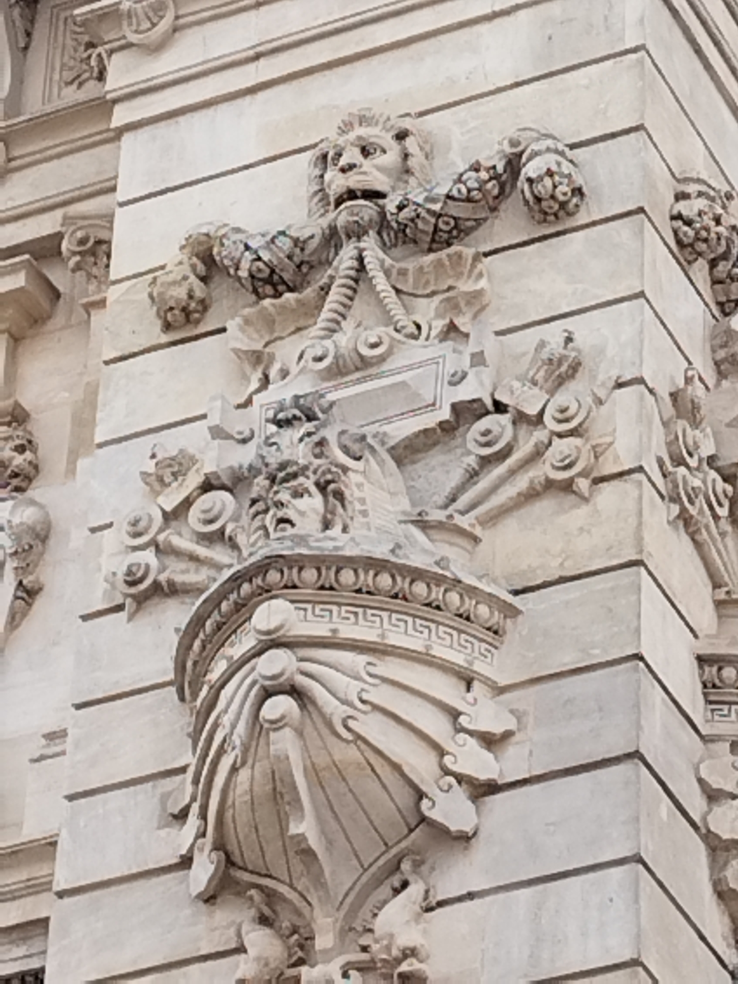 The image shows a close-up of intricate stone carvings on the exterior of a building, likely a historical or architectural landmark. The carvings depict mythological figures, including a lion's head with flowing mane and a human-like face with a distressed expression, possibly a gorgon. There are also decorative elements such as rosettes, acanthus leaves, and geometric patterns. The stonework is detailed and appears weathered, suggesting age. The lighting suggests it is daytime, with shadows indicating the direction of the sun. This facade detail is located on the former Royal Palace in Bucharest, Romania.
