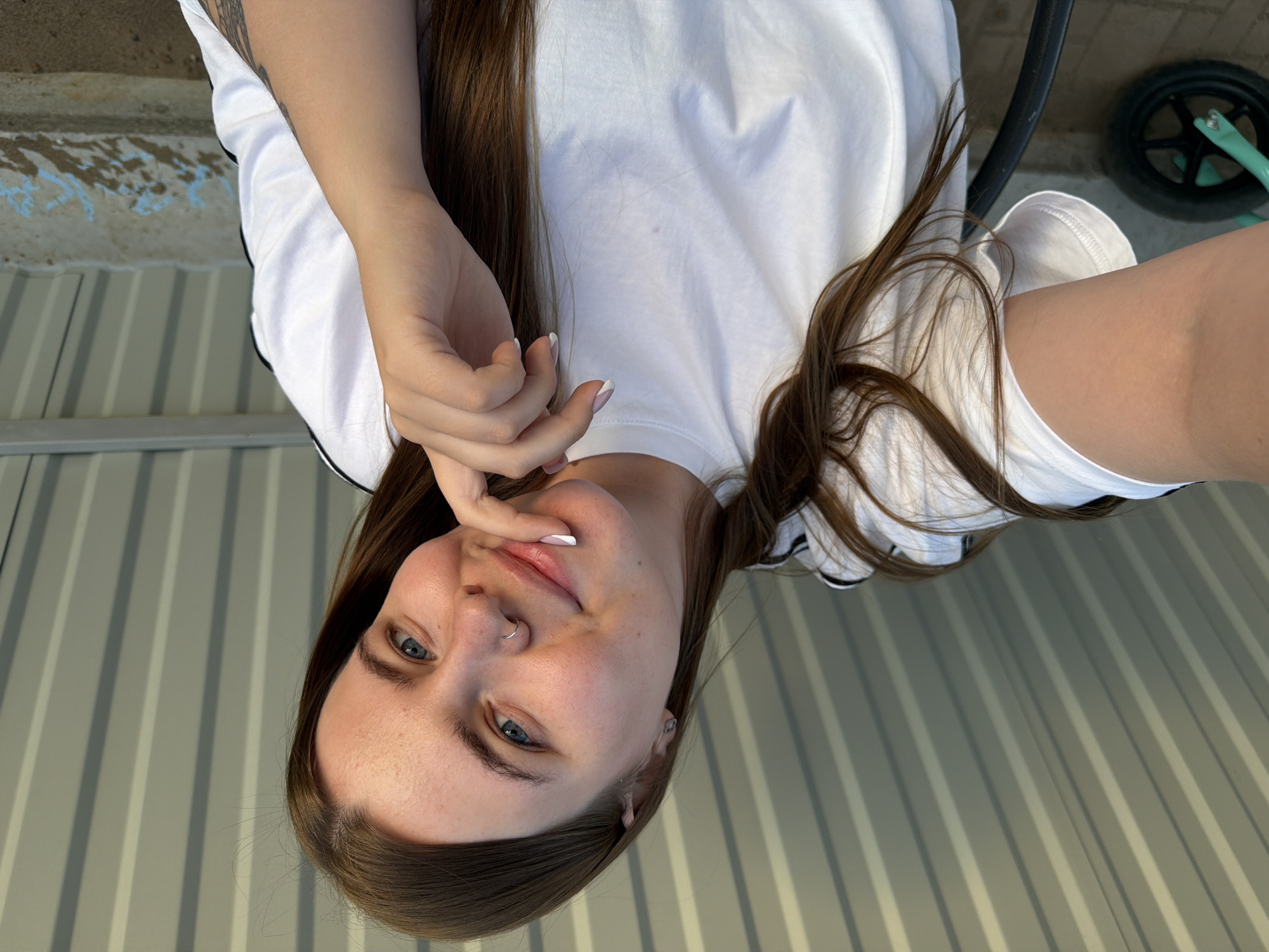 The image shows a young woman with light skin and blue eyes, wearing a white t-shirt with black trim. She has long brown hair. Her hand is near her face, with her finger touching her lip. She has a nose ring and what appears to be an earring. The background consists of a striped metal surface, part of a building, and a tire. The location is Chaikovsky, Russia.