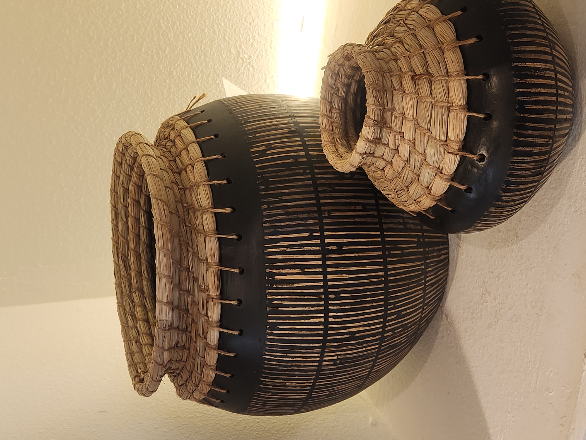 Two decorative baskets are displayed on a white, textured surface. The baskets are made of woven natural fibers and dark material, possibly clay or painted ceramic. Both baskets feature a wider body that tapers towards the bottom, with a woven fiber rim and lid. The dark portions of the baskets are decorated with vertical lines etched into the surface. The larger basket is positioned behind and to the left of the smaller one. The background is a textured, off-white wall, with the scene illuminated by soft lighting from the right, casting shadows. The location is identified as the City of Maitland, United States of America.