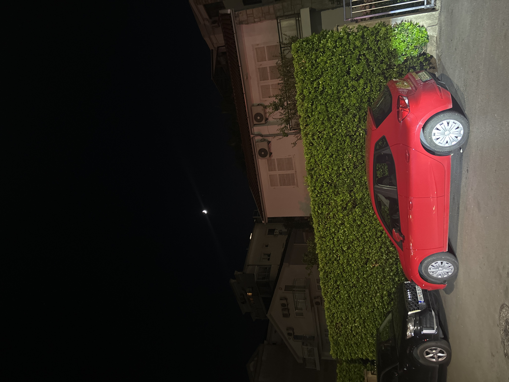 The image shows a nighttime scene on a street in Budva, Montenegro, with a red hatchback and a black SUV parked next to a dense green hedge. The red car is parked on the right side of the frame, while the black car is on the left. Buildings with illuminated windows are visible behind the hedge, and a crescent moon is in the dark sky. The street surface is asphalt, with a manhole cover visible in the foreground. The scene is illuminated by artificial lighting, casting shadows and highlighting the vehicles and the hedge.
