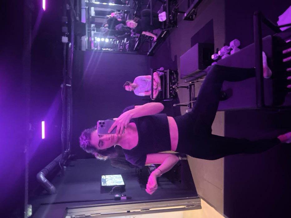 A woman in workout attire is taking a selfie with a phone. She is in a workout studio with other people and equipment visible in the background. The lighting is primarily purple. The woman is wearing a black crop top and black leggings. The equipment in the background includes what appears to be a reformer machine and dumbbells. One person is working out on a piece of equipment directly behind the woman taking the picture.