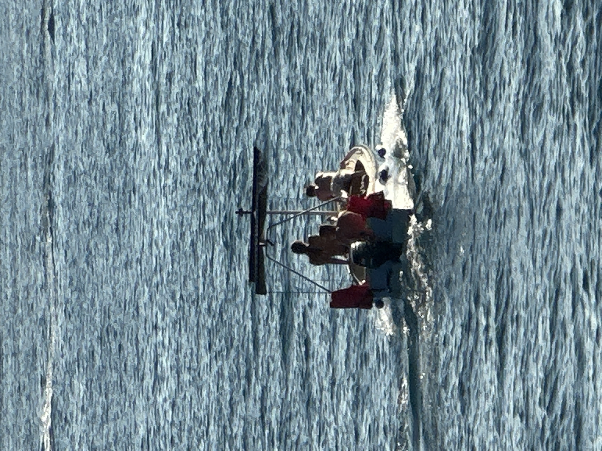 The image shows a top-down view of a small boat with several people on it, sailing on a body of water that appears to be a lake or a calm sea. The water has a textured, rippling surface, with streaks of light reflecting off it, suggesting it is either sunny or the water is turbulent. The boat is white with a dark mast and appears to have a sail partially unfurled. There are at least five people visible, some wearing red and blue clothing. They are positioned around the center of the boat, some appearing to be actively engaged in sailing, while others are seated. The overall impression is of a leisurely activity on the water.
The scene is bathed in natural light, and the water's surface texture implies it is daytime. There are no clear indicators of the specific time of day, weather conditions beyond the appearance of the water, or specific emotions on the faces of the individuals. The location is identified as Budva, Montenegro, which is a coastal town known for its beaches and historical sites, suggesting this could be a view of the Adriatic Sea. The image is a high-angle shot, providing a unique perspective of the boat and its occupants against the vast expanse of the water. There is no visible text in the image.