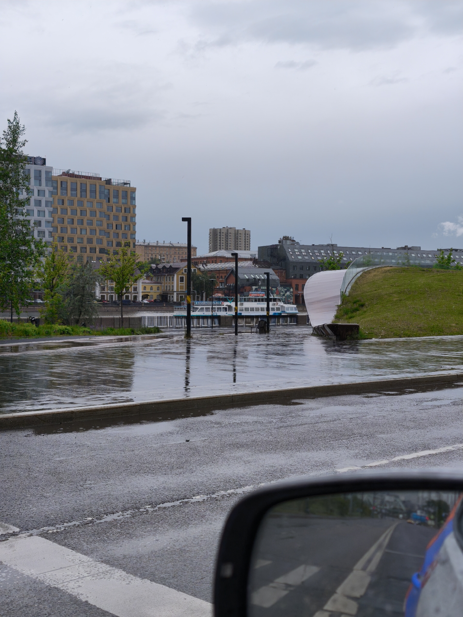 The image shows a rainy urban scene in Moscow, Russia. In the background, a modern building with a gridded facade stands next to traditional buildings. A pedestrian zone flooded with water stretches along the embankment, reflecting the buildings and street lights. Several boats are docked at the river. A landscaped hill with a modern pavilion is visible on the right. In the foreground, a wet road with a crosswalk leads to the embankment. A car's side mirror is visible at the bottom right corner. The sky is overcast, suggesting a continuation of the rain.