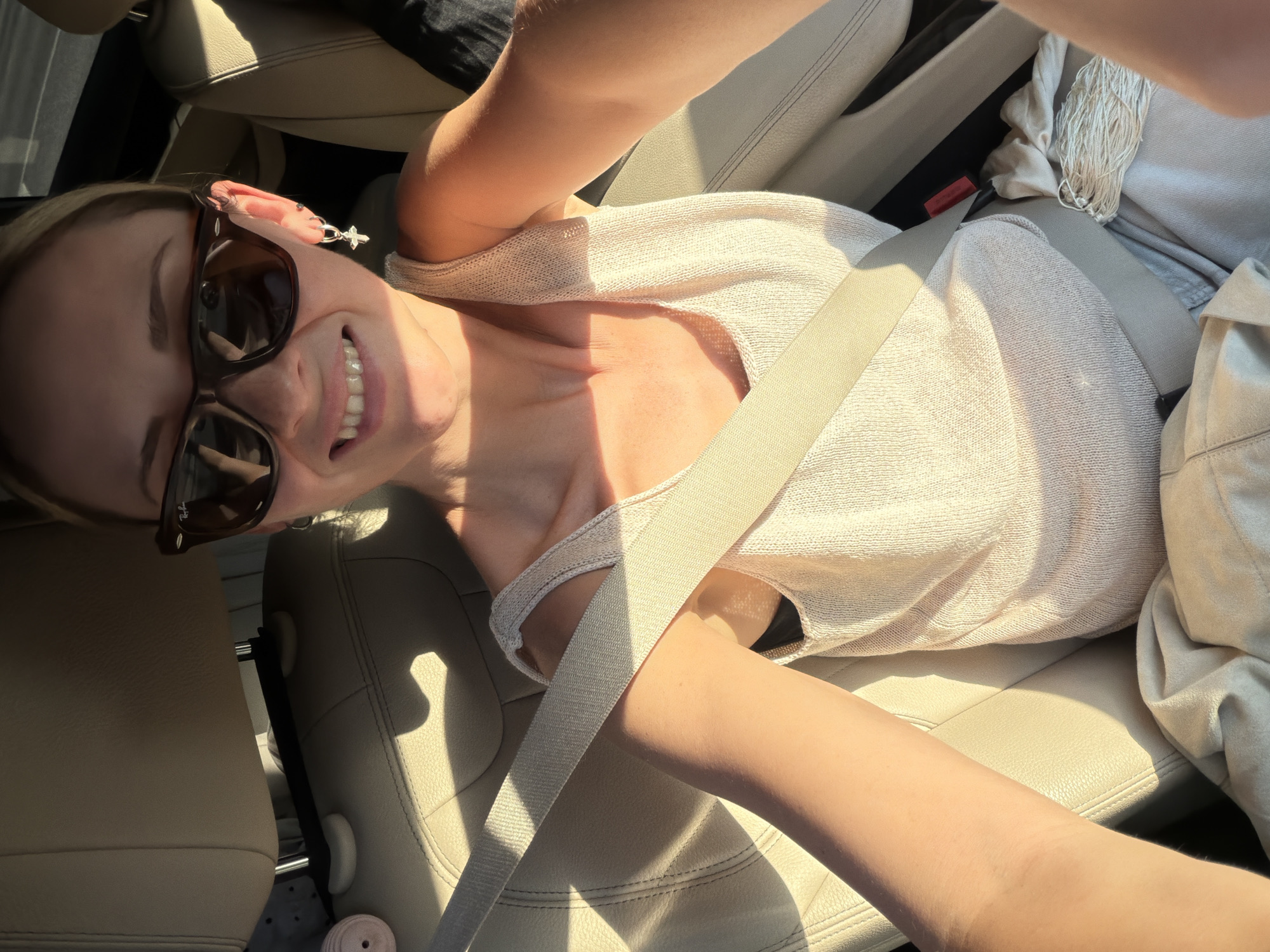 The image displays an adult female occupant of a vehicle, taking a selfie. The scene is brightly lit by direct sunlight, creating strong shadows within the car's interior. The woman, identified by her blonde hair, is wearing dark-tinted sunglasses, which feature a visible "Ray-Ban" logo on the right lens. She is smiling, revealing her teeth. Her attire consists of a light-colored, possibly beige or off-white, knitted tank top. A light-colored seatbelt is fastened across her chest and lap. On her visible right ear, she wears a silver-toned hoop earring with a dangling cross charm, accompanied by a smaller stud earring higher on the earlobe.
The vehicle's interior features light-colored (beige or tan) leather upholstery on the front passenger seat and a section of the rear seat. A light-colored bundle of strings or yarn is present on the front passenger seat. A dark-colored fabric item is observed on the floorboard adjacent to the passenger seat. The specific geographical context for this scene is the city of Khimki, Russia.