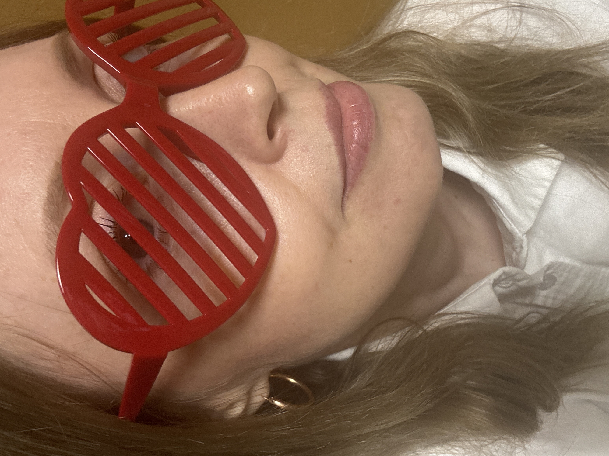 A close-up shot depicts a person with light skin and blonde hair, wearing red slatted sunglasses that partially obscure the left eye. The person is lying down, with a white collared shirt visible at the neck. The location is unknown and cannot be determined from the image. The image primarily focuses on the person's facial features, including the nose and lips, and there are no clear actions or interactions occurring in the frame. The environment is generic, with a gold or yellow-toned background visible at the top of the frame. The subject is positioned in a way that implies relaxation or repose. There is no other information that can be derived from the image.