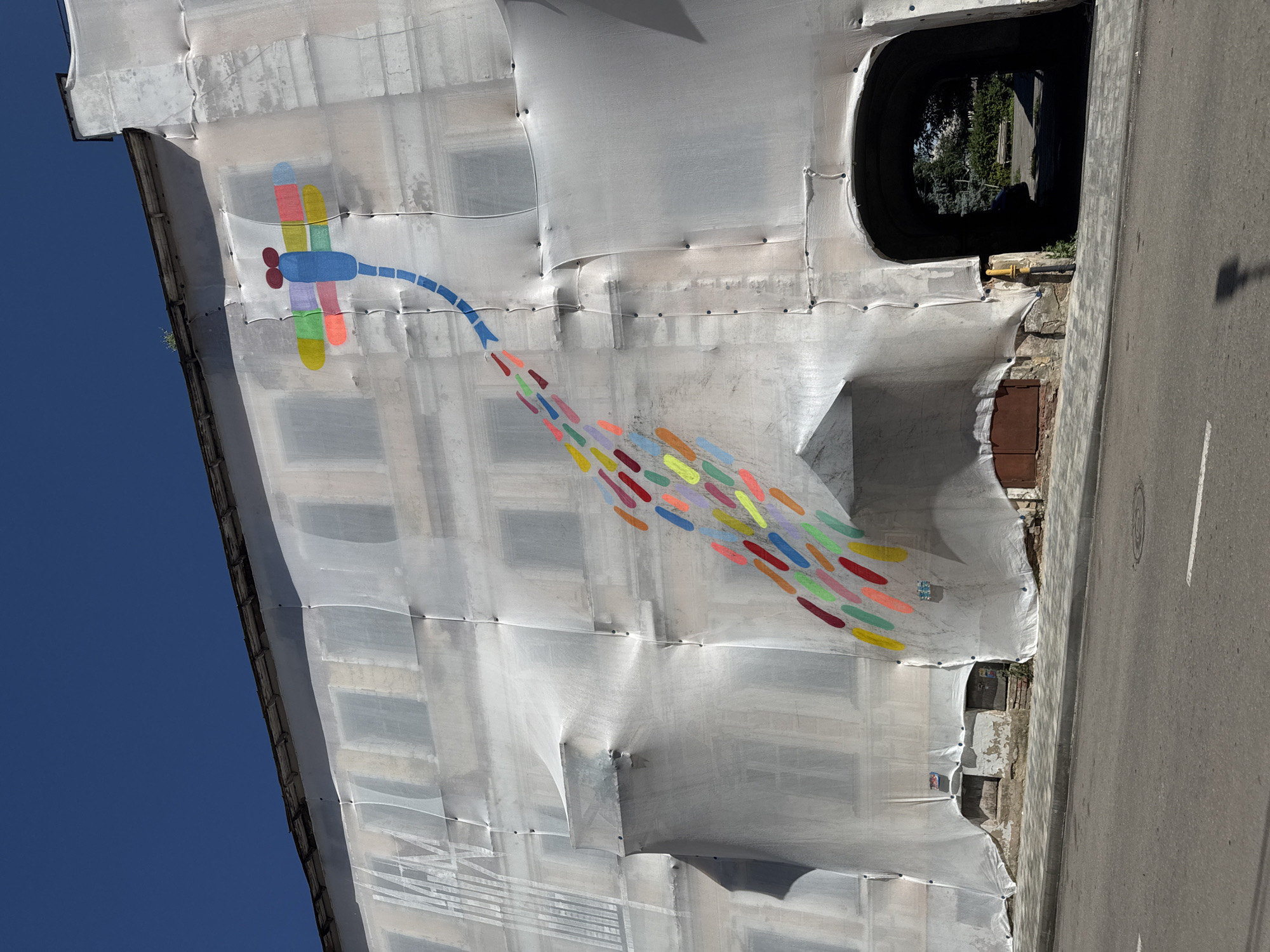 A street art mural of a colorful dragonfly with a trail of colored droplets extends across a building facade covered in white scaffolding sheeting in Yelets, Russia. The dragonfly, depicted in blue, red, yellow, green, and pink, is positioned at the top left of the building. Its body is blue, with red eyes and wings made of segmented colored rectangles. A trailing tail of numerous small, elongated droplets in various bright colors—including red, orange, yellow, green, light blue, and purple—extends downwards and to the right, creating a dynamic visual effect. The building itself appears to be under renovation or construction, as indicated by the extensive sheeting. The sheeting is draped over the windows and architectural details, creating folds and shadows. Below the main facade, there is an arched entryway, and the street in front is paved with asphalt and bordered by a sidewalk made of stone pavers. The sky is clear and blue.