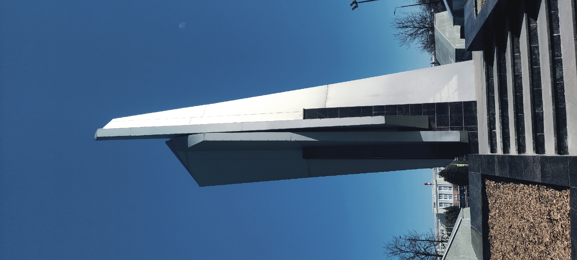 The image shows a monument in Fryazino, Russia. The monument features an angular, white structure with dark grey and black accents, set against a bright blue sky. The monument is adjacent to a street with buildings visible in the background. In the foreground, there is a textured surface, possibly a stone or gravel area. Some bare tree branches are visible. A streetlight pole can be seen in the background as well.