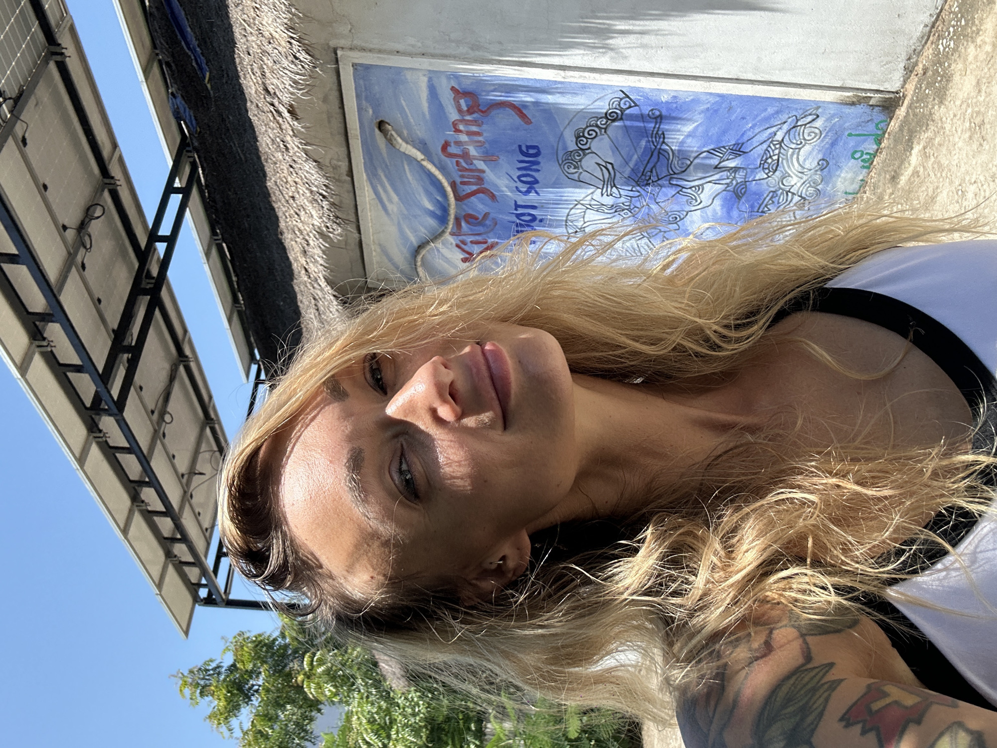 In this close-up, outdoor, selfie-style photograph, a woman with blonde, wavy hair and tattoos on her left arm is positioned in the foreground, facing slightly to the right. She is wearing a white tank top with black trim. Behind her, a building with a thatched roof is visible, with a blue mural painted on its side. The mural depicts a rope snake and text that appears to read "Kite Surfing" and "LOT SONG." Above the building, solar panels are mounted on a metal frame. The environment suggests a sunny day, with a clear blue sky. The building, with its thatched roof and mural, is characteristic of some establishments found in the coastal tourist areas of Mũi Né, Vietnam, a popular destination for kitesurfing.