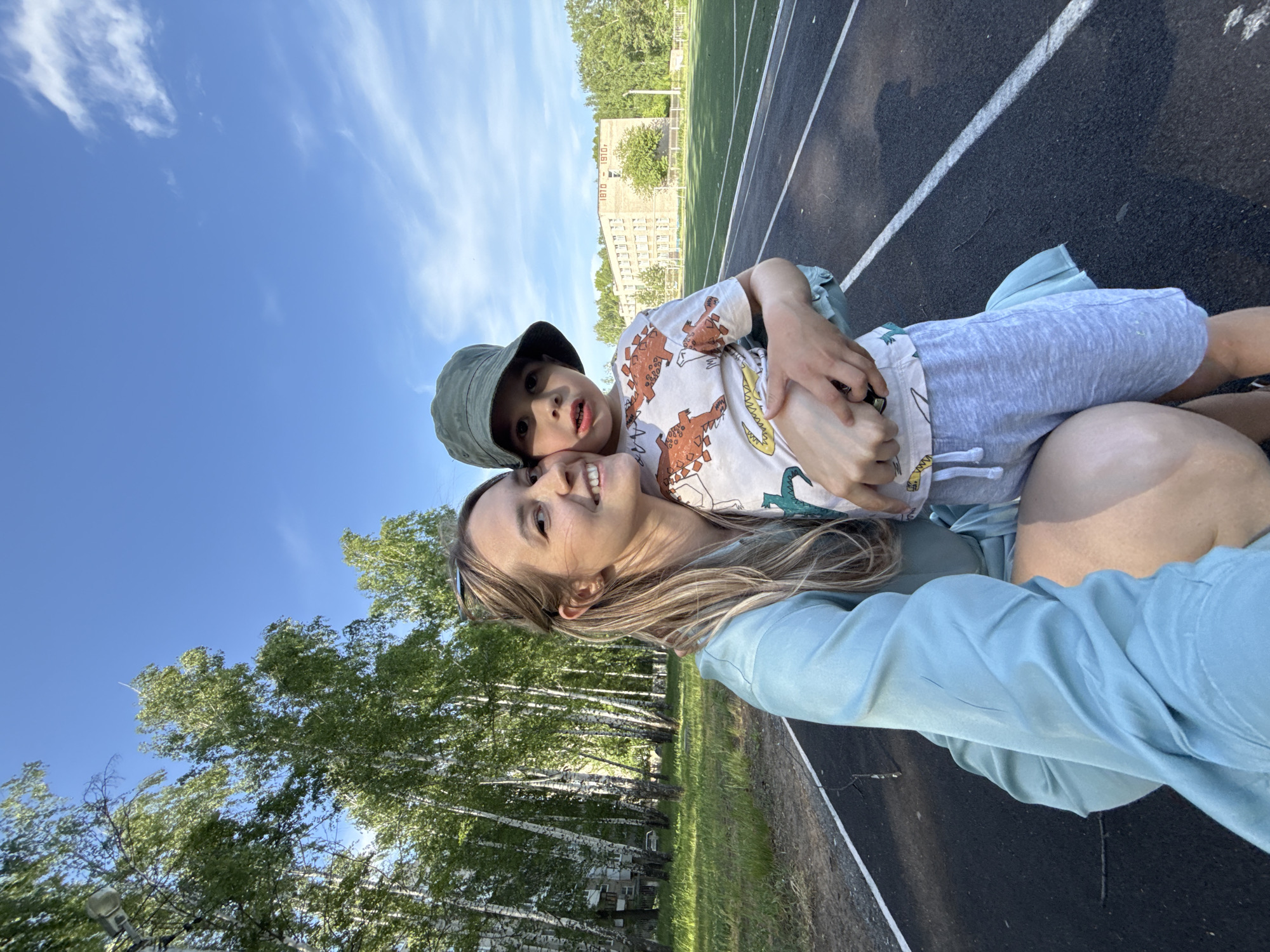 A mother and child are posing for a selfie on a running track. The mother is fair-skinned with long, light brown hair and is wearing sunglasses and a light blue shirt. The child, also fair-skinned, is wearing a light-colored shirt with dinosaur prints, gray shorts, and a dark green bucket hat. The child is being held by the mother. The background includes a bright blue sky with some clouds, trees, a building with "1810 - 1910" on its side, and a section of a running track with black asphalt and white lines. The location is in the city of Chaikovsky, Russia.