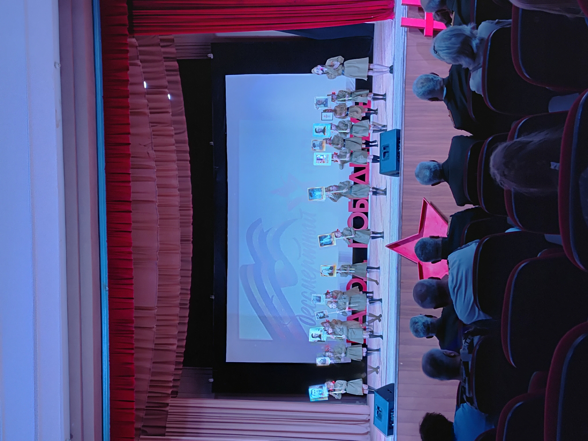 The image shows a theater performance taking place in an unknown location. Children in uniform are performing on stage, holding up pictures. Behind them is a screen displaying a logo. The audience is seated in red chairs. Red curtains adorn the sides of the stage. A red star is also visible near the stage. The lighting appears to be theatrical with spotlights on the performers.