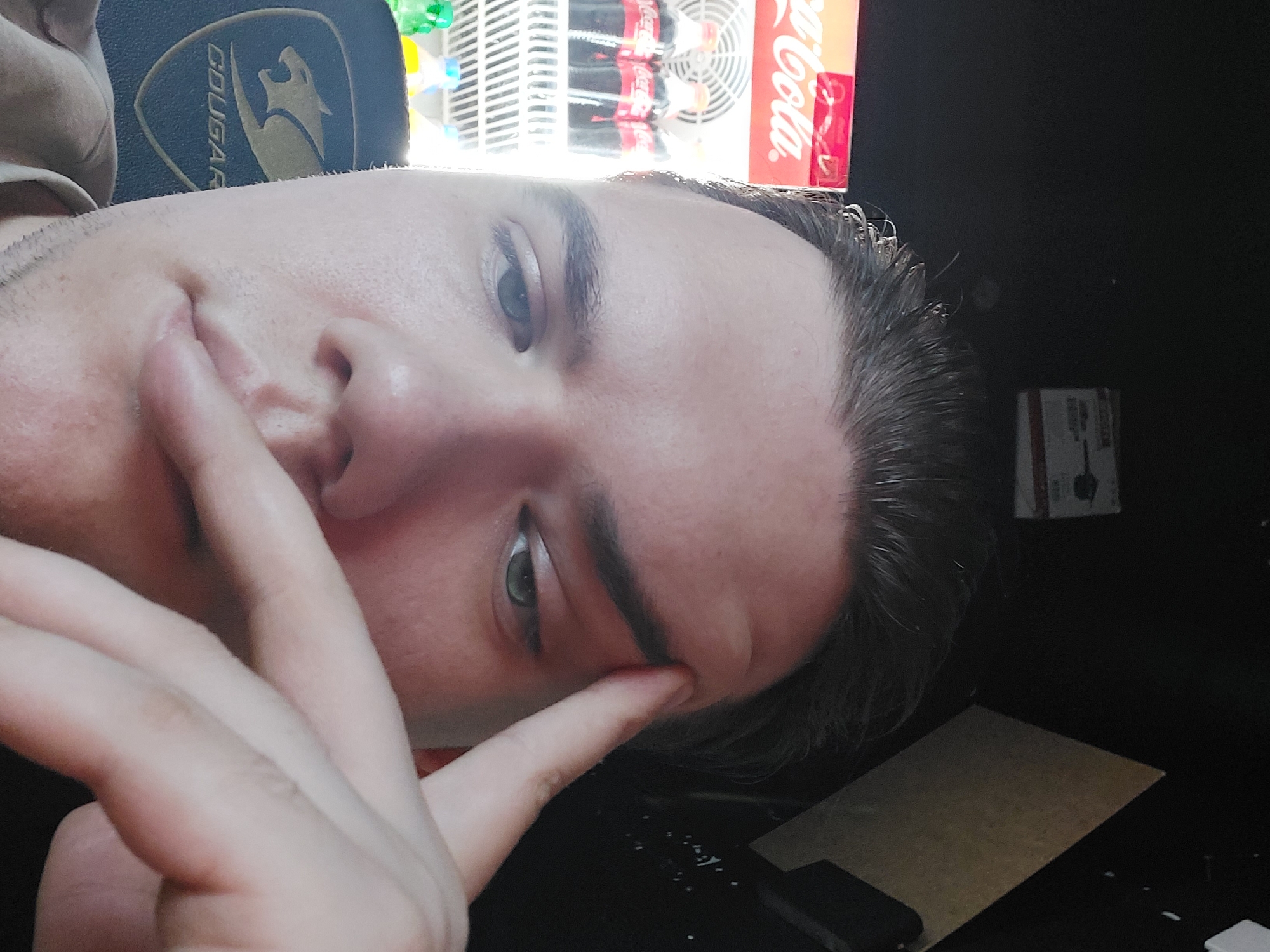 A man's face is prominently featured in the image. He is partially obscuring his face with his hand, touching his lips and forehead. He is wearing a hat with a "Cougar" logo. Behind him, a Coca-Cola refrigerator is visible, suggesting a store or market setting. The environment is dark, with a cardboard box and a phone on a table or surface. The location is unknown, and there is no verifiable information about Tashkent, Uzbekistan.
