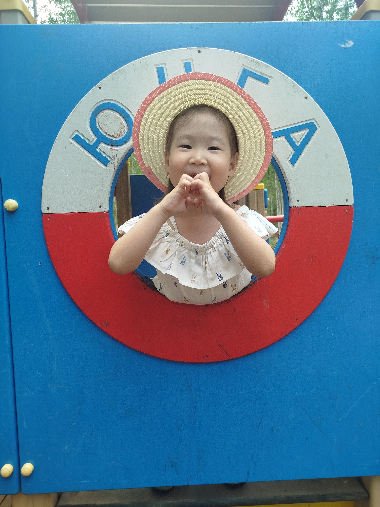 A young East Asian girl wearing a straw hat with a pink trim is peeking through a circular opening in a blue playground structure. The opening has a white and red border with blue lettering visible. The girl is smiling and making a heart shape with her fingers near her mouth. She is wearing a white top with a subtle pattern of bunnies. The background appears to be an outdoor playground with trees visible through the opening. The lower part of the image shows the blue structure with yellow knobs on the left side. The location is identified as Mirny, Russia.