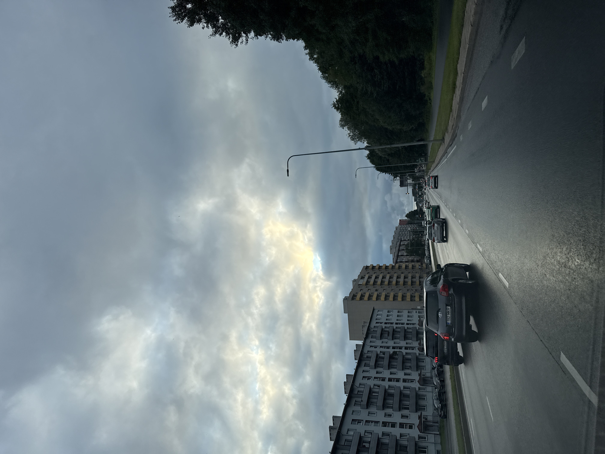 The image captures a view of a multi-lane road in Tallinn, Estonia, likely from inside a vehicle. The sky is overcast with dramatic clouds, and a patch of sunlight breaks through on the left side, illuminating the clouds. On the left side of the road, there are several multi-story apartment buildings with balconies. On the right side of the road, a dense line of trees runs along the pavement. Several cars are visible on the road, driving in the same direction as the implied viewpoint. One dark-colored car in the foreground has a license plate visible. Streetlights are present along the roadside. The road surface appears wet, suggesting recent rain. The overall impression is a typical urban street scene under a moody sky.