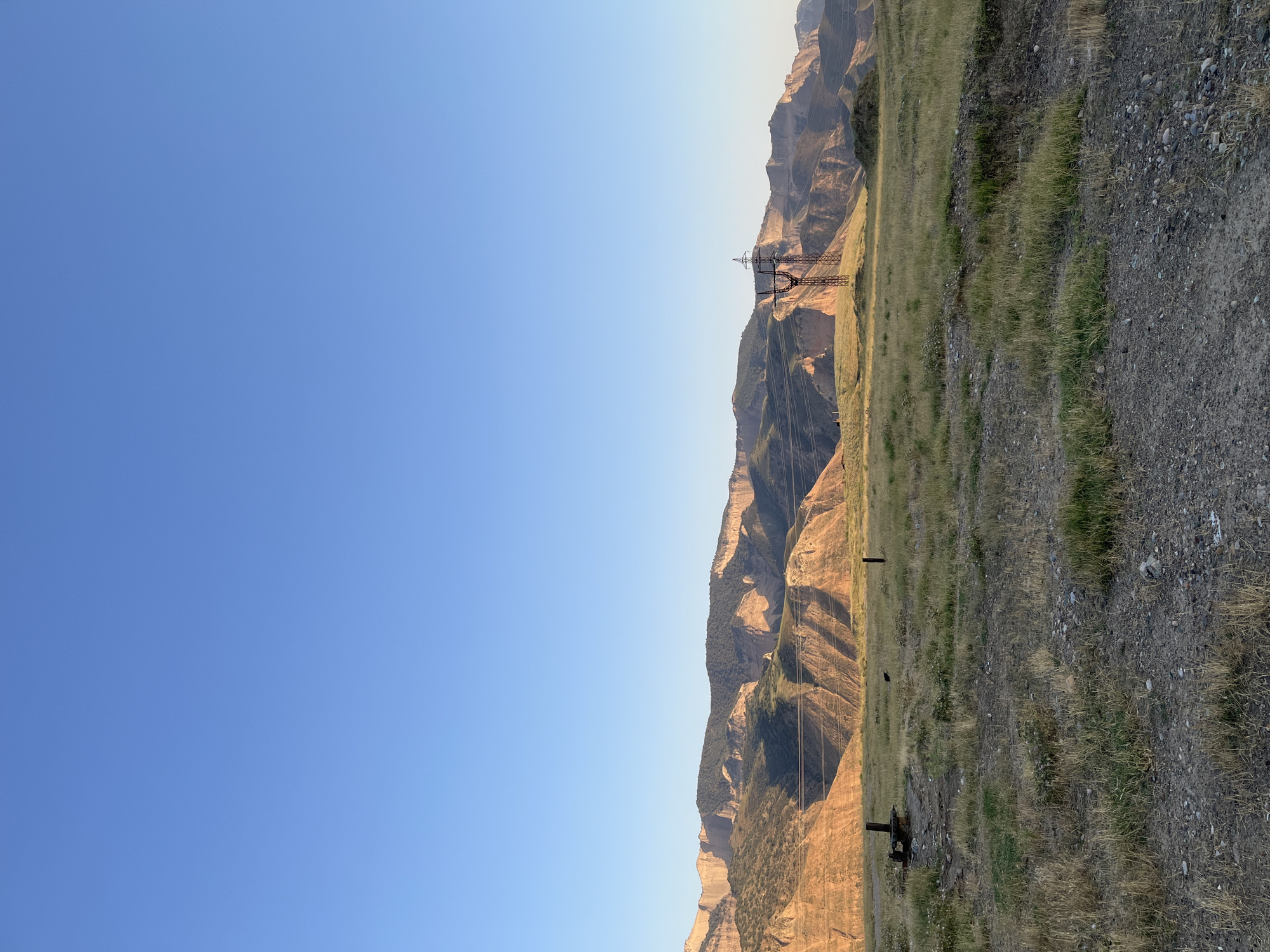 The image depicts a vast, open landscape in the city of Mailuu-Suu, Kyrgyzstan, under a clear, bright blue sky. In the foreground, a dry, grassy field stretches out, with patches of dirt and gravel visible. A few weathered wooden posts are scattered across the field, and to the left, a rusty metal object resembling old machinery or debris rests on the ground.
Dominating the middle ground and background are undulating hills and mountains, their layered rock formations illuminated by the warm glow of sunlight, casting shadows that highlight their rugged texture. The slopes are covered in sparse vegetation, with darker green patches suggesting more concentrated plant growth.
A prominent feature in the middle ground is a tall, skeletal electricity pylon with wires stretching across the frame. The pylon, made of dark metal, stands starkly against the natural backdrop. The overall scene conveys a sense of remoteness and natural beauty, with hints of human industrial presence in the form of the pylon and debris. The lighting suggests it is either early morning or late afternoon.
