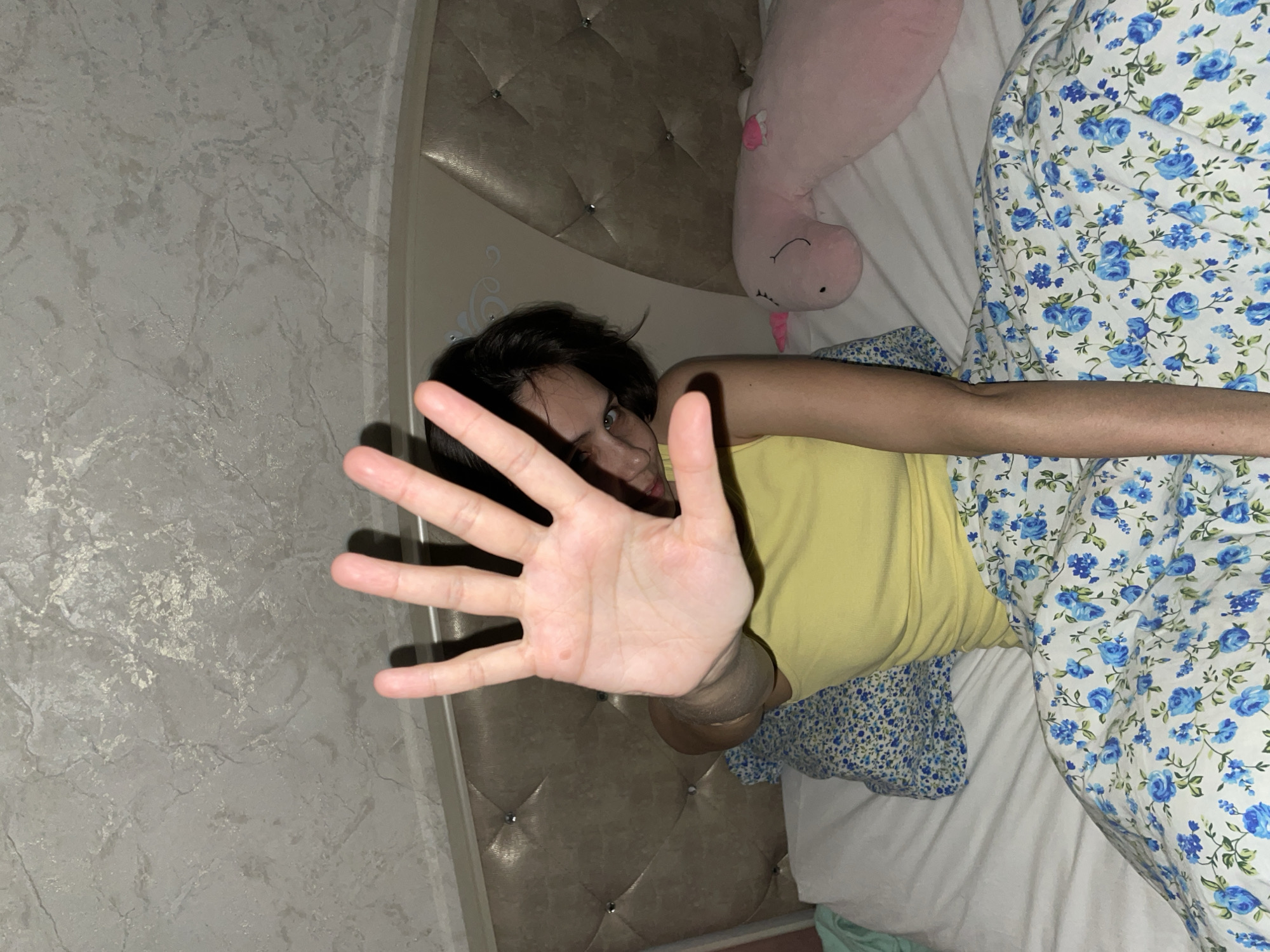 A person lies on a bed with floral-patterned sheets and a small pillow with a similar floral design. A pink stuffed unicorn is positioned near the head of the bed. The person is wearing a yellow tank top and extends a hand towards the camera. The headboard is padded and studded. The wall next to the bed is textured in a gray pattern. The location is not discernible from the image.