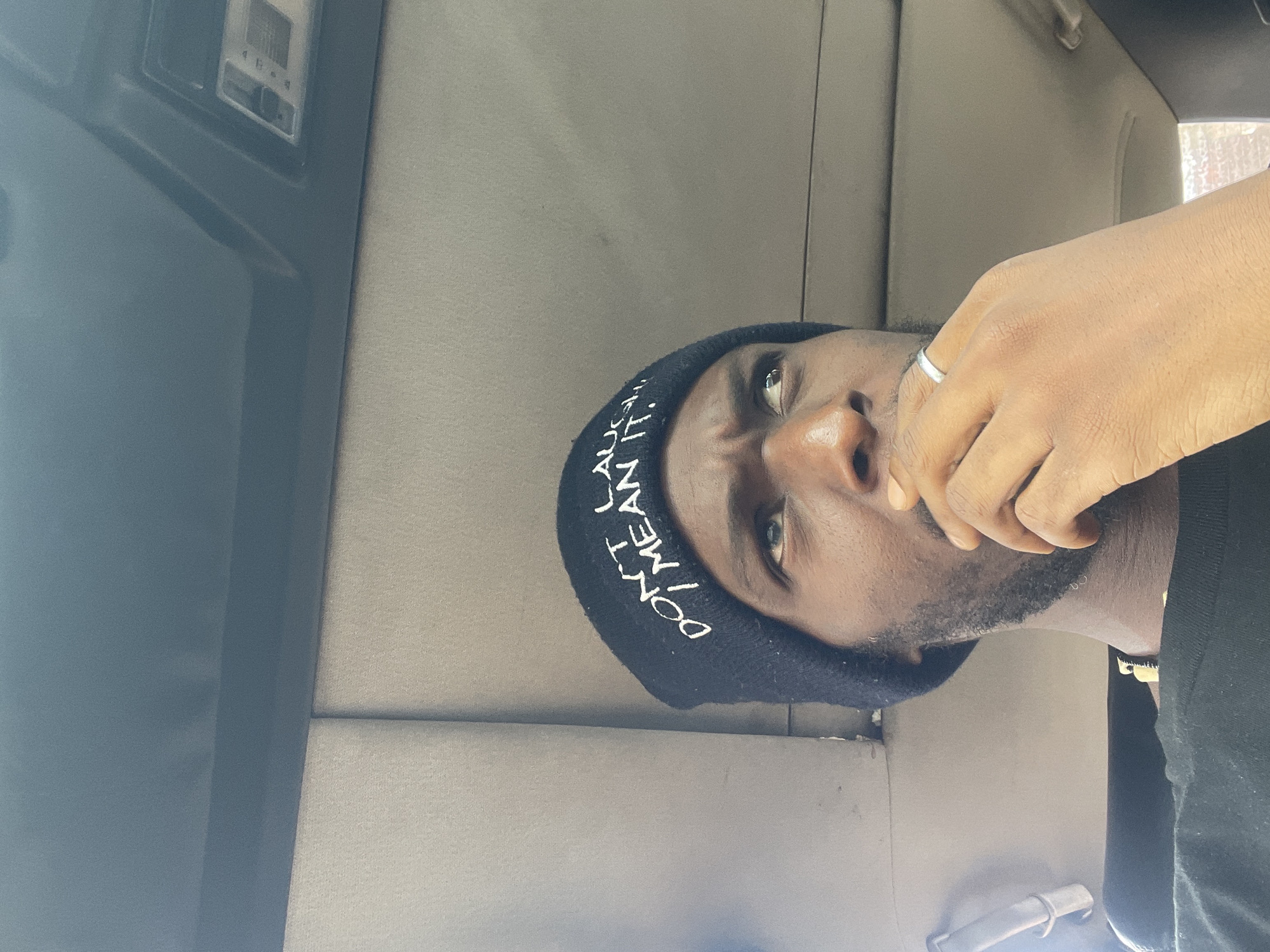 A mid-angle, eye-level shot shows a Black male with a goatee wearing a black beanie, seated inside a vehicle, likely in Jos, Nigeria. His right hand is partially obscuring his mouth. The interior of the vehicle includes gray upholstery and a black dashboard. The light suggests daytime.
