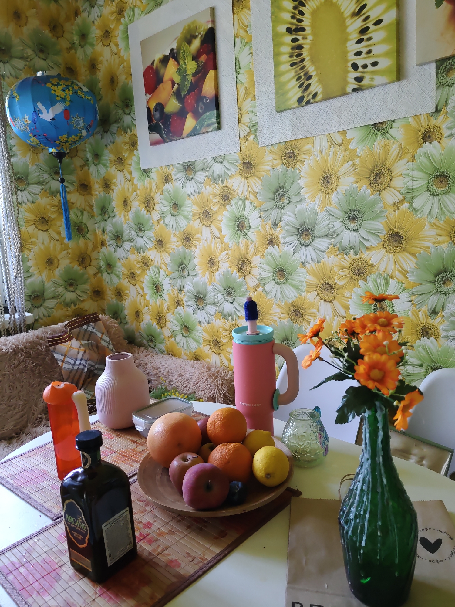 The image depicts a still life scene on a table with a floral wallpaper background. In the foreground, a dark bottle with a label that reads "ARCANE MAURITIUS ISLAND" is positioned on a bamboo placemat. To the left of the bottle are two orange containers and a white cylindrical object. A wooden bowl filled with fruits, including oranges, lemons, and apples, is centrally located. Behind the fruit bowl, a pink water bottle with a straw is visible. To the right of the water bottle, there is a small, decorative glass jar. Further to the right, a bouquet of orange flowers is arranged in a dark green, wax-dripped bottle. Two paper bags are placed behind the green bottle. The wall is adorned with a bright floral wallpaper featuring yellow and green flowers, and two framed pictures hang on it. One picture shows a fruit salad, and the other displays a slice of kiwi fruit. A blue Chinese lantern with a white bird design hangs from the upper left corner of the image. The scene appears to be set indoors. The provided information states that this image is located in the city of Rostov-on-Don, Russia.
