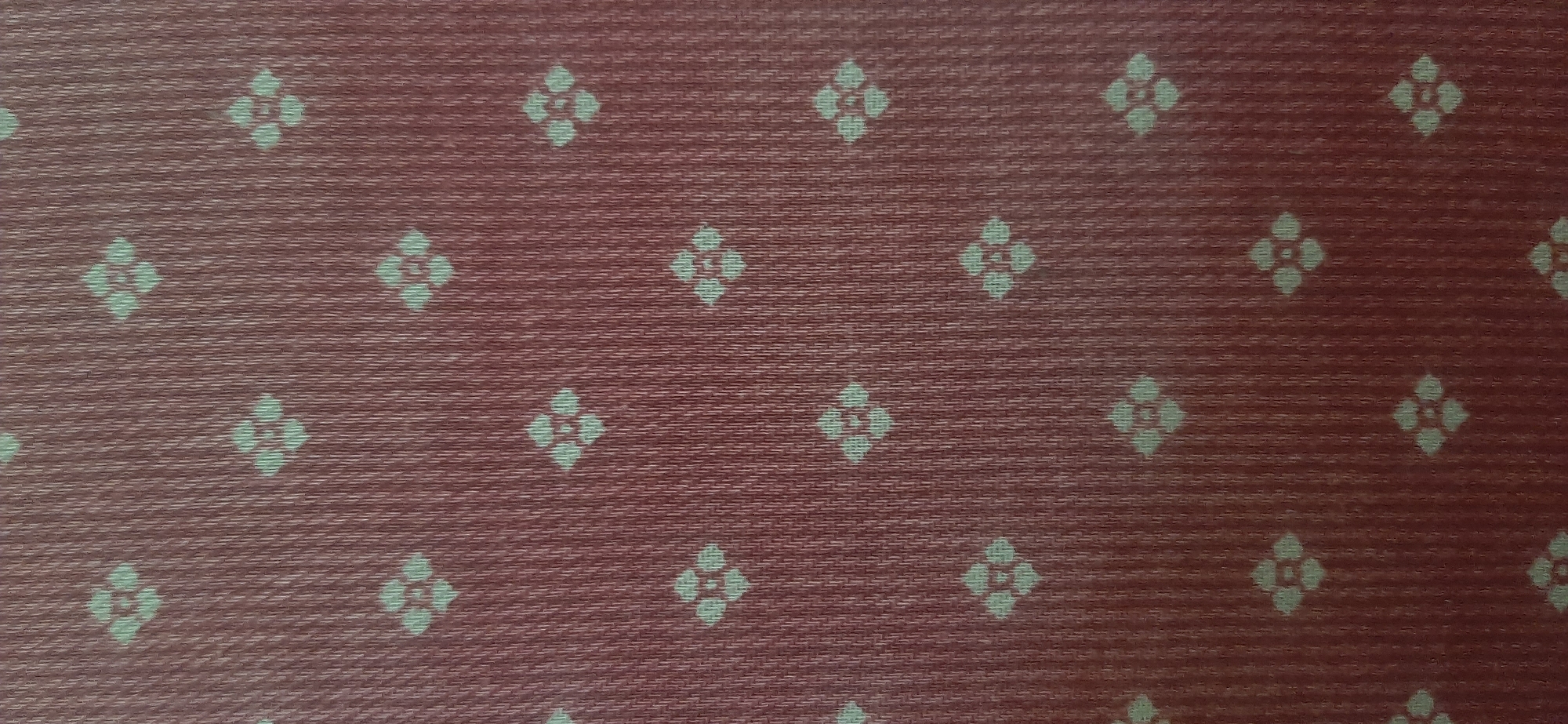 The image is a close-up, overhead view of a textured, reddish-brown fabric with a repeating pattern of small, light-colored floral motifs. The fabric appears to be woven, with visible horizontal and diagonal threads creating a textured surface. The floral shapes are stylized, resembling four-petal flowers, each arranged in a diamond orientation within a square grid. The lighting is even, and there are no shadows, suggesting it was taken in a well-lit indoor environment or under diffuse natural light. There is no visible text, people, or activity. The image focuses solely on the pattern and texture of the fabric.